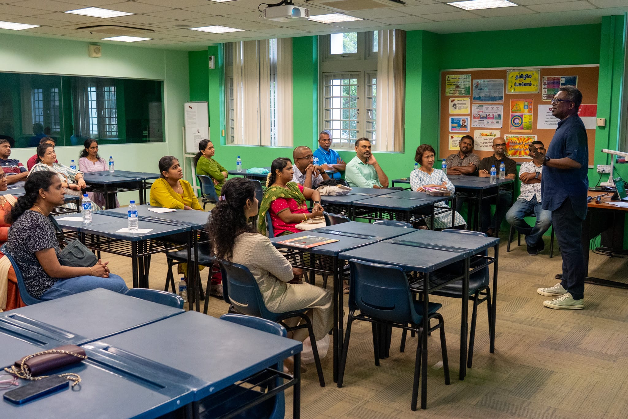 Man stands at front of green classroom speaking to seated adults at desks with water bottles and posters on wall.