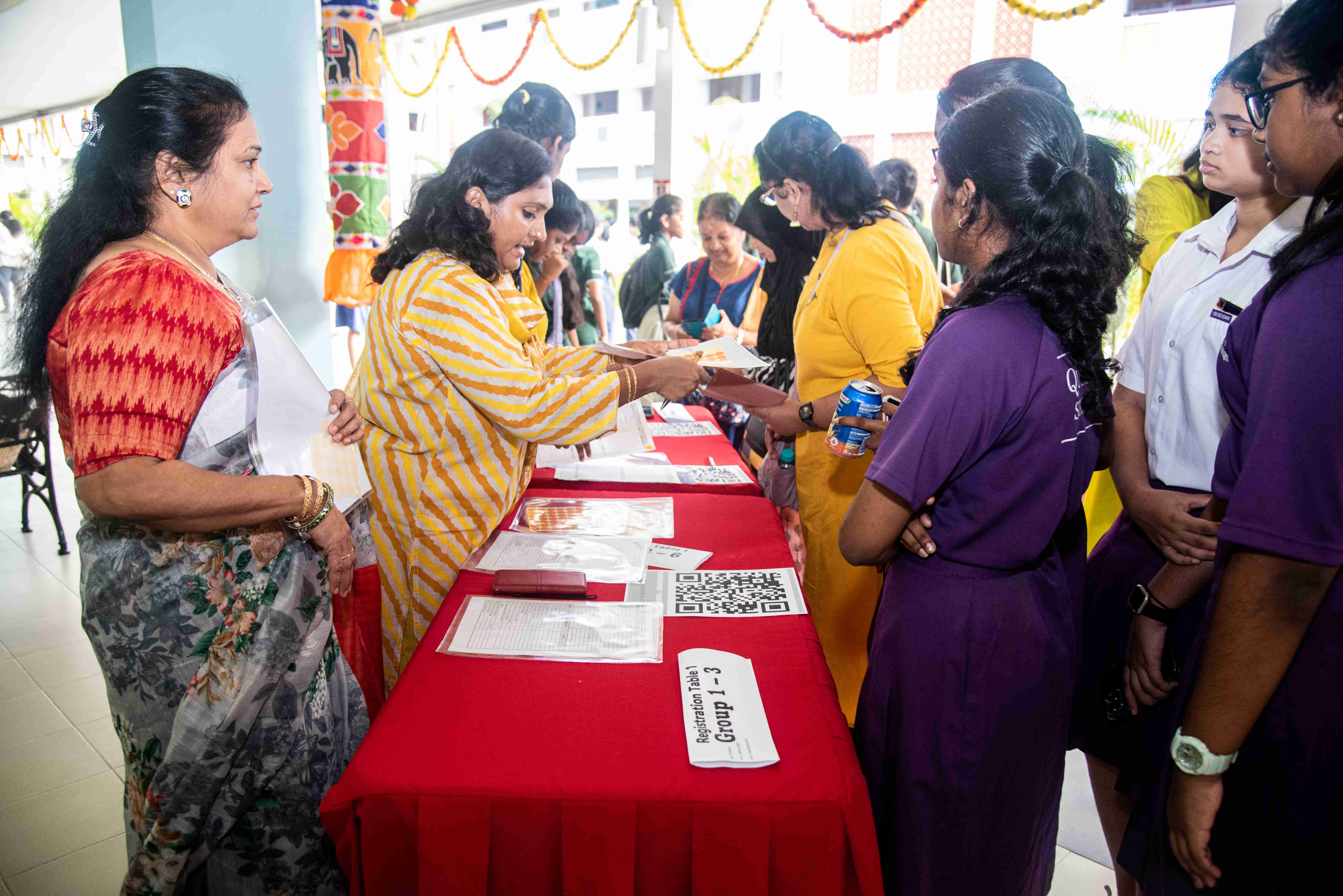 Women at a red registration table handing papers to students in purple uniforms; QR code and forms on table.