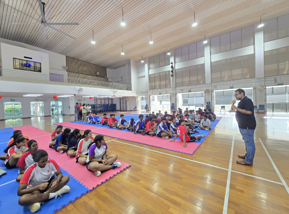 Man speaks into a microphone as students sit on blue and pink mats in a large indoor gymnasium.