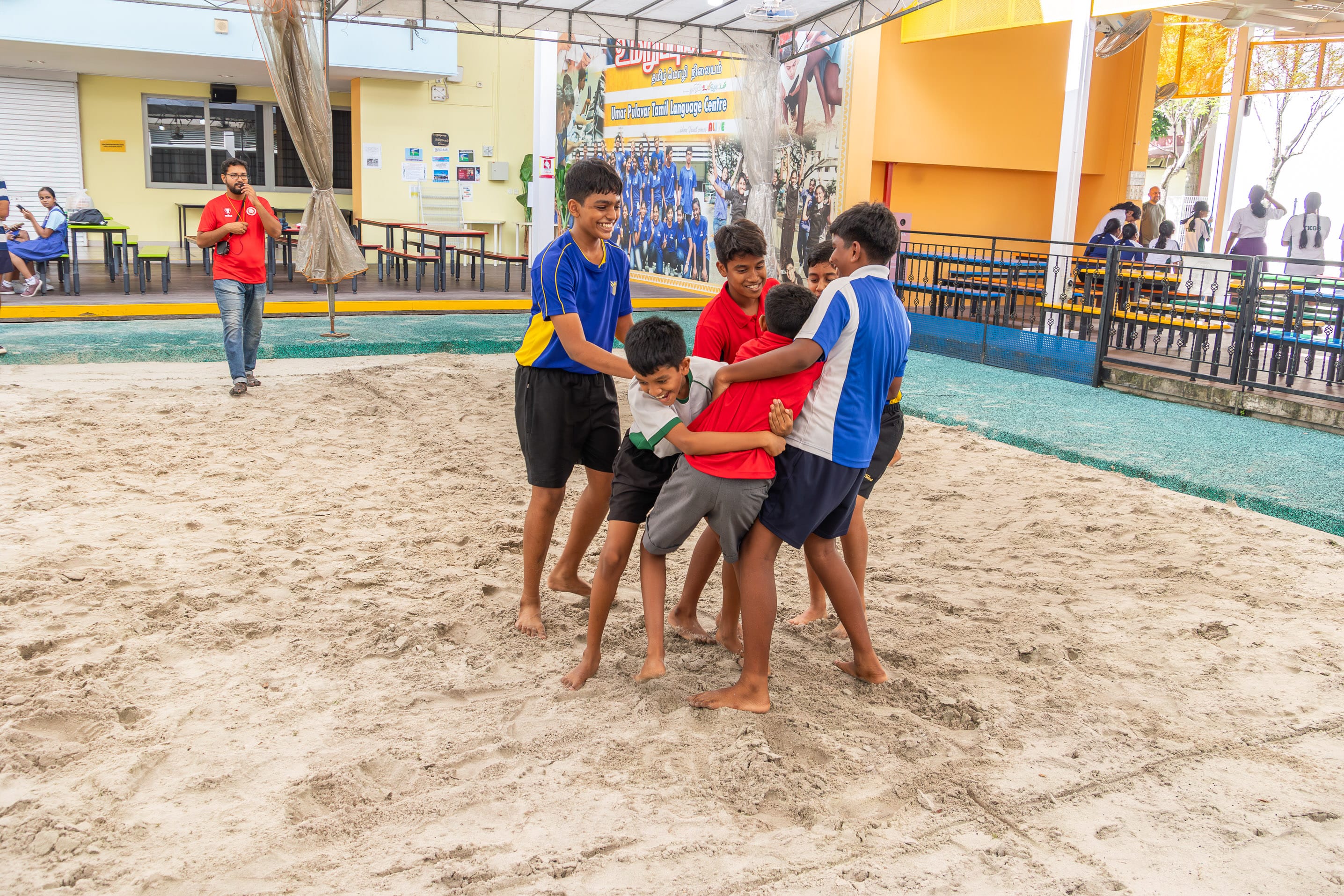 Boys in sports jerseys playfully grapple in a sand pit while others watch in a school courtyard.