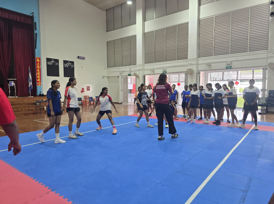 Students on blue mats in a gym watch a coach in a maroon shirt while two girls demonstrate a move.