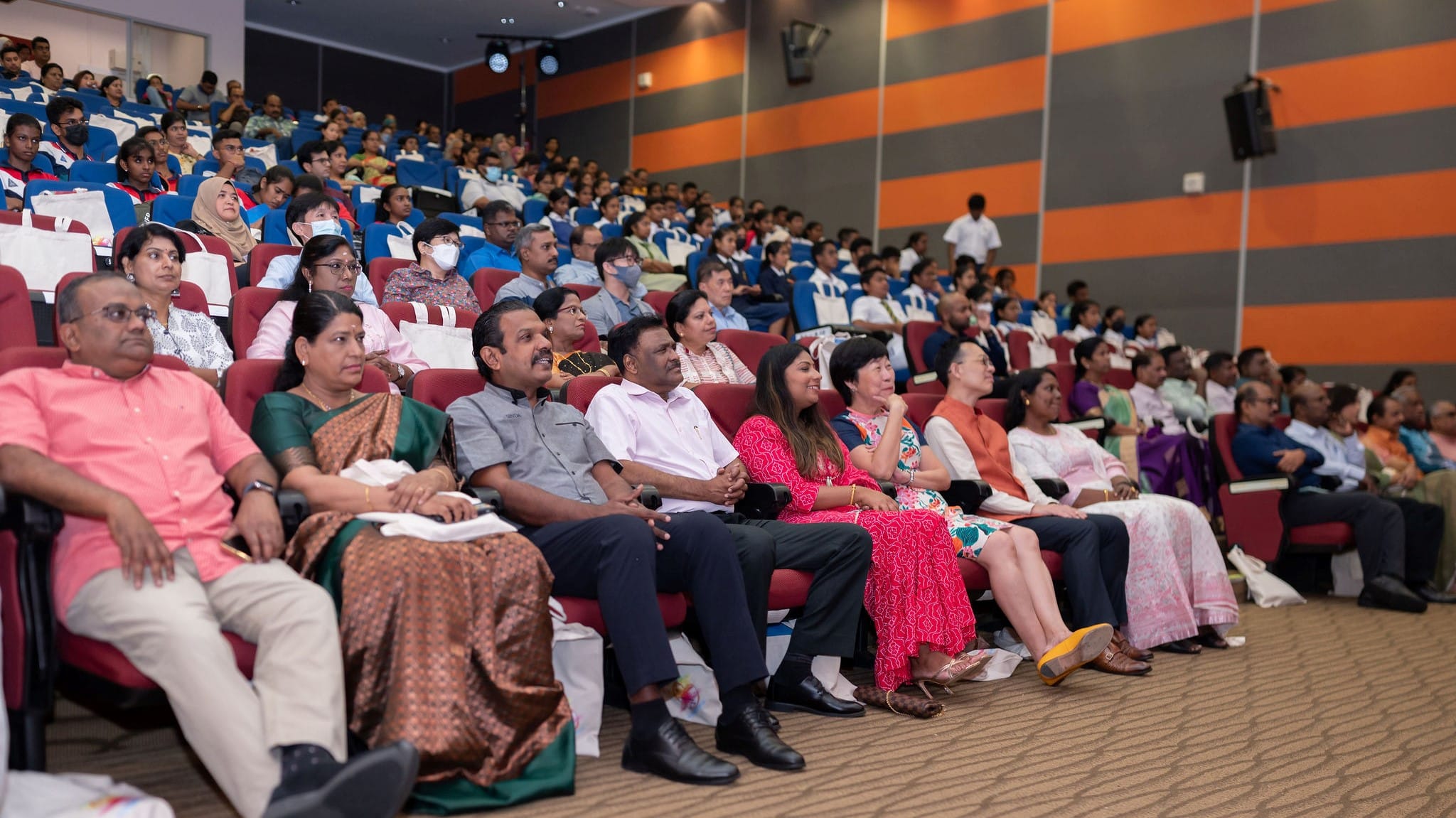 Audience seated in a large auditorium with red chairs, facing forward; many students in uniform behind.