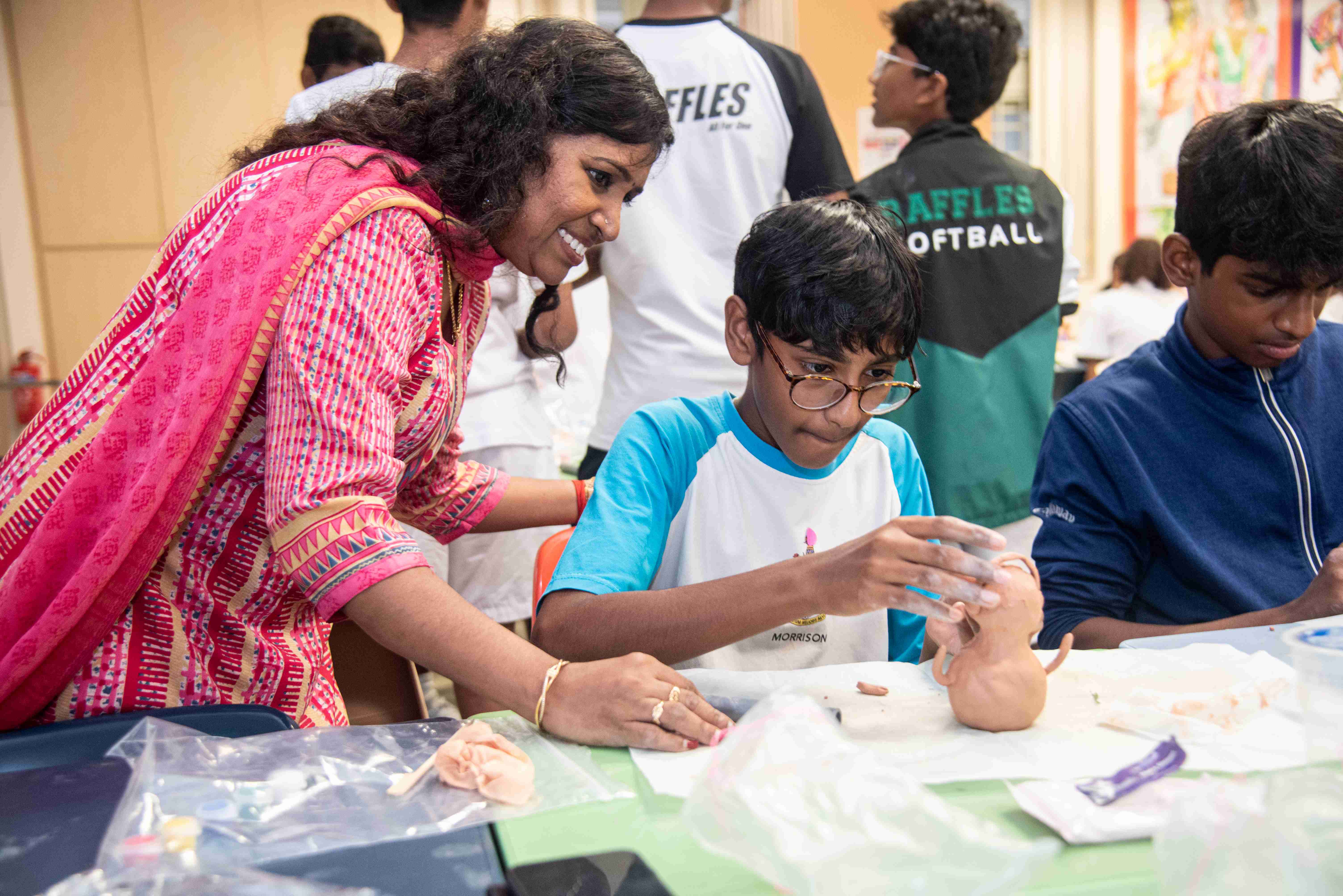 Woman leans over a boy with glasses working on a model ear at a table, with other students nearby.