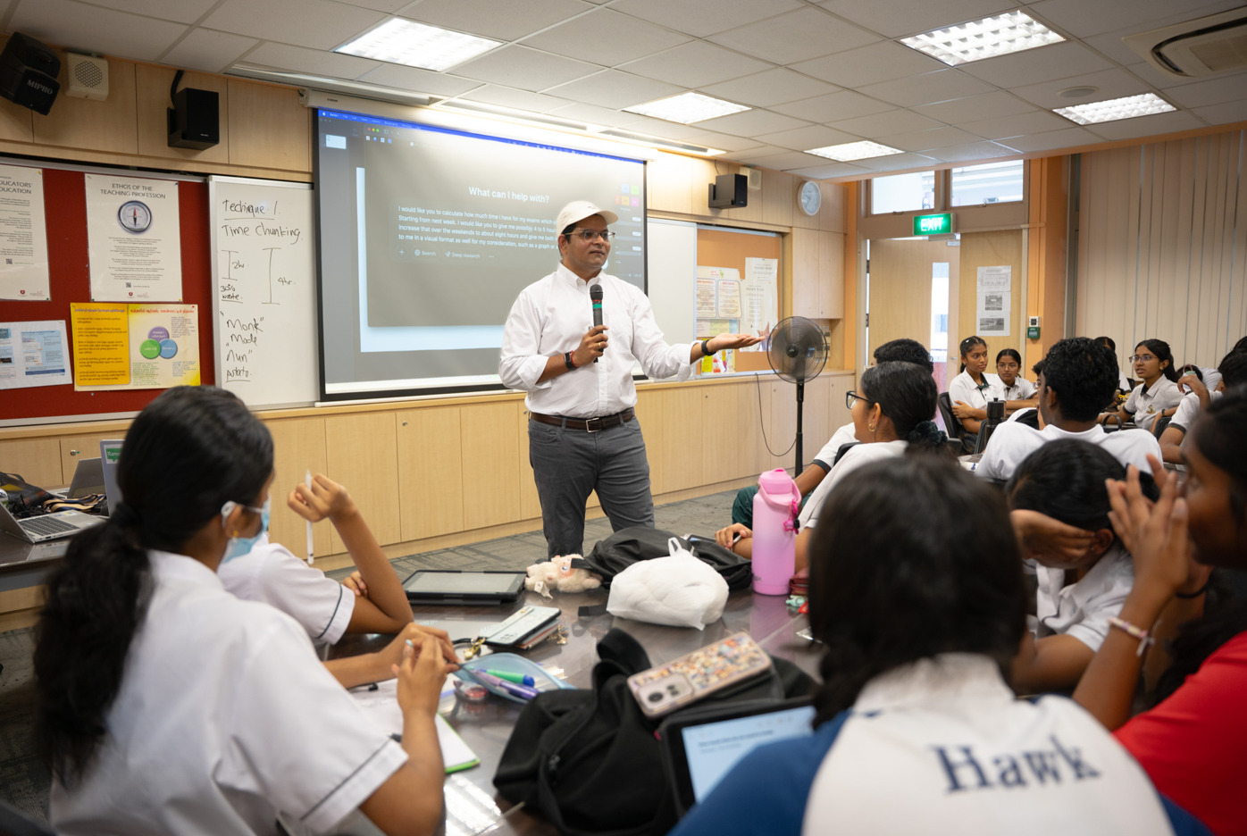 Man with microphone speaks to students in a classroom, with a projected screen and whiteboard behind him.