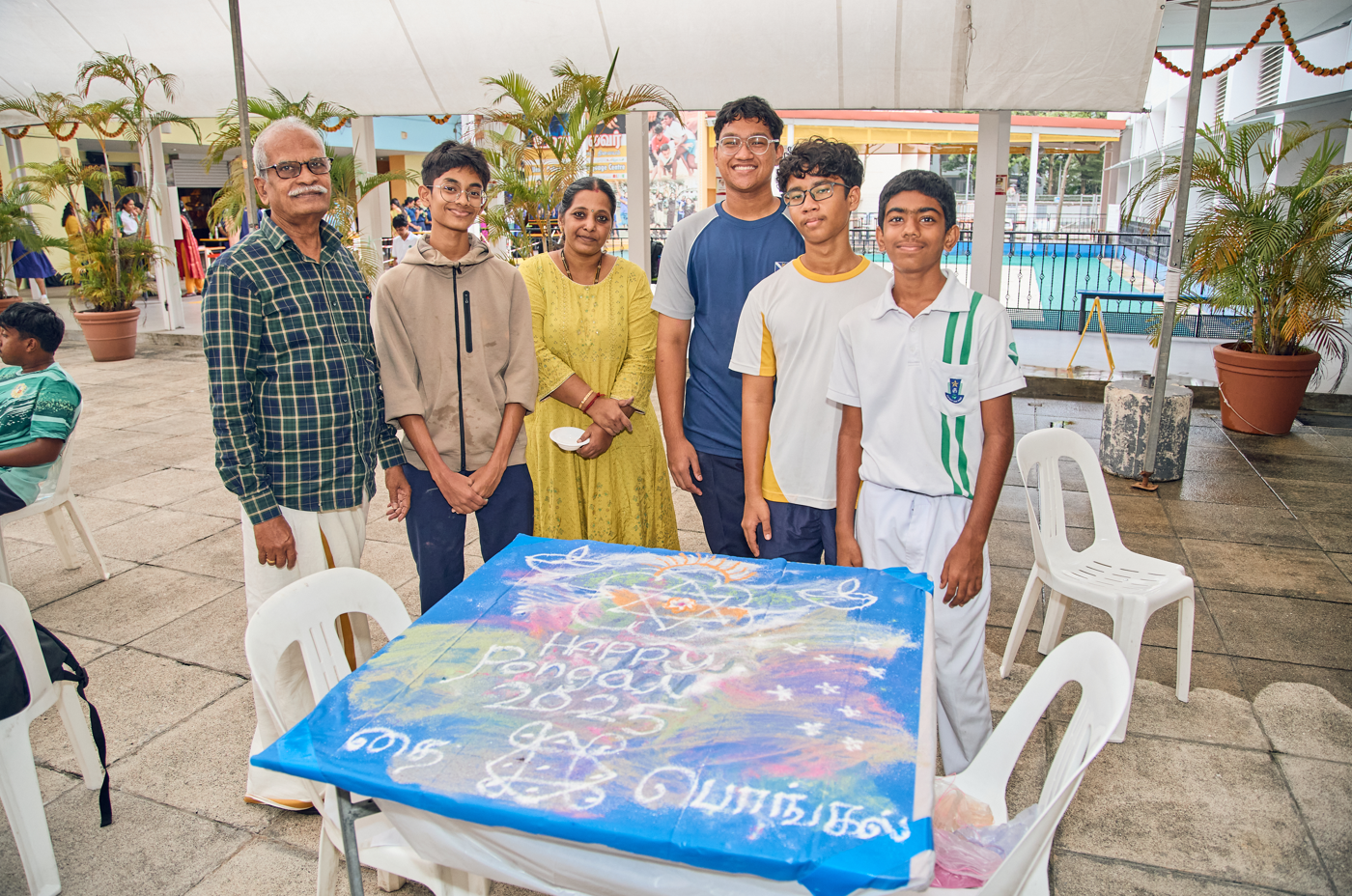 Six people pose by a blue chalk-art table under a canopy, with chairs, plants, and a pool behind.