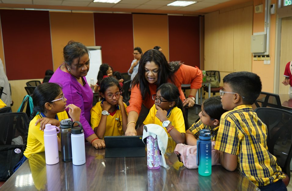 Two women guide schoolchildren in yellow uniforms around a tablet on a table with water bottles.