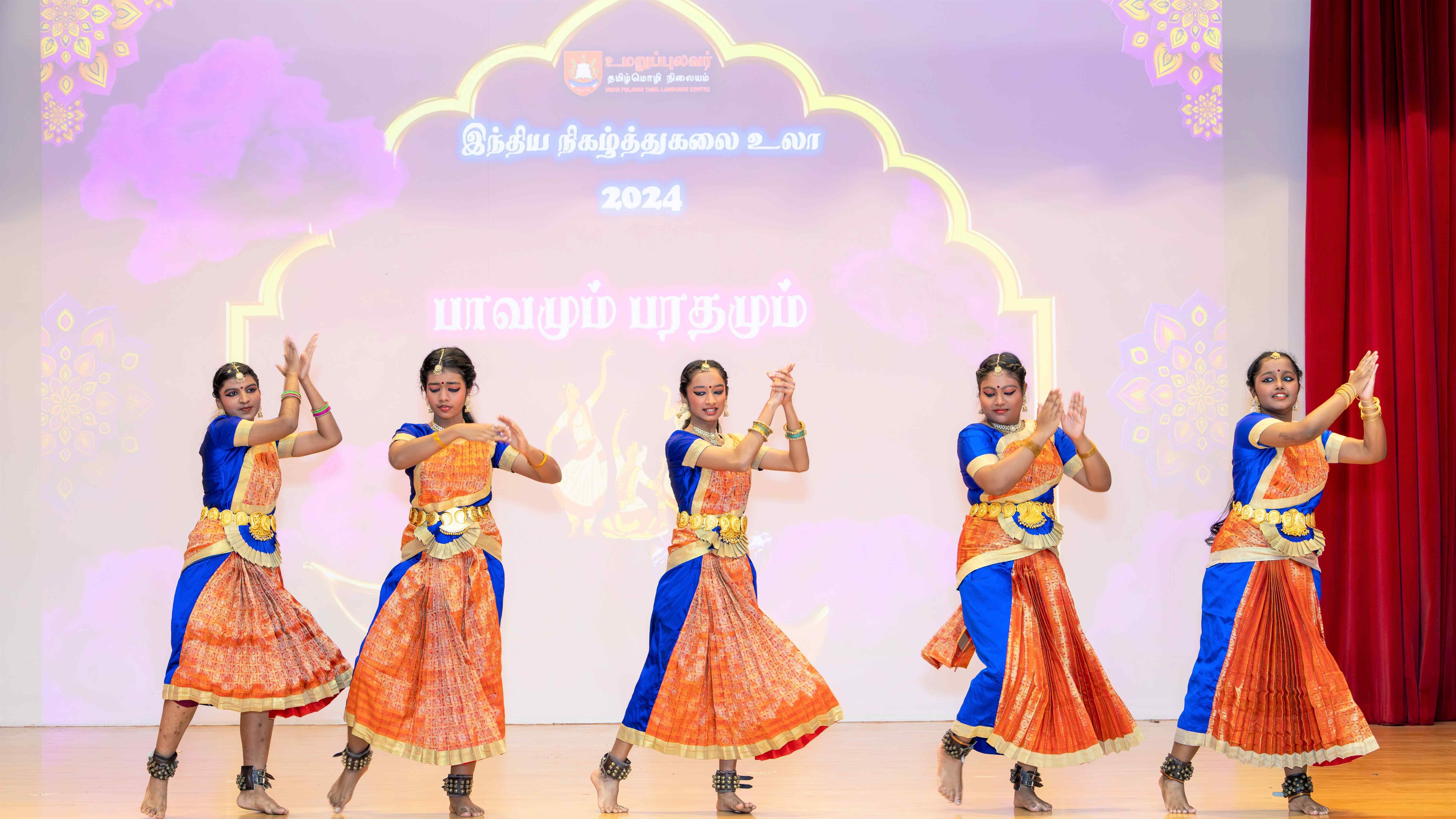 Five dancers in blue and orange costumes perform on stage before a pastel backdrop with Tamil text.
