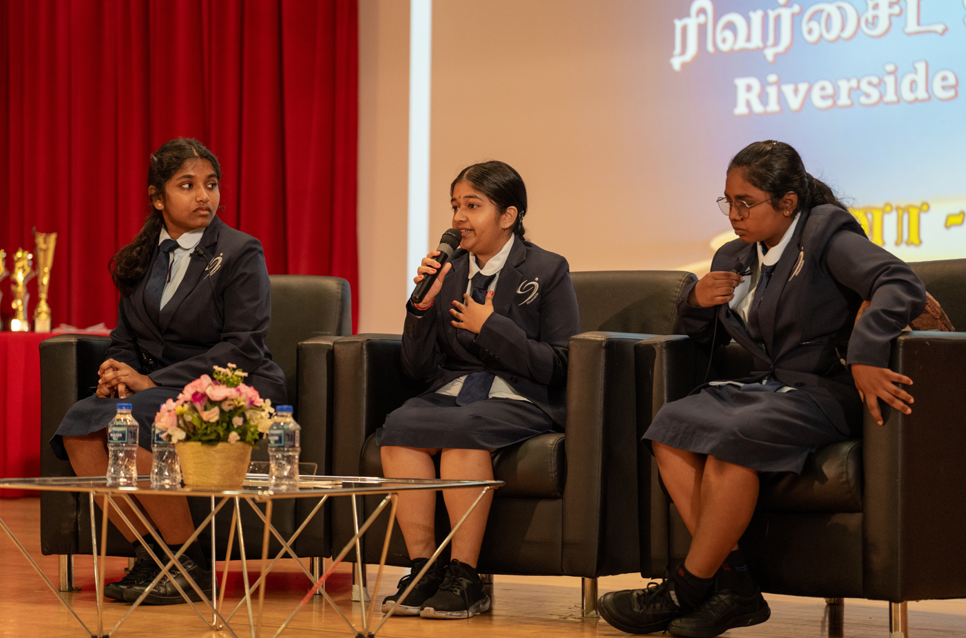 Three students in school uniforms sit on black chairs onstage; center student speaks into a microphone.