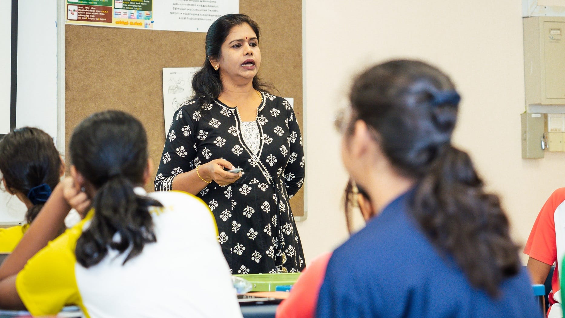 Teacher in patterned dress speaks to seated students in a classroom, holding a remote near a bulletin board.