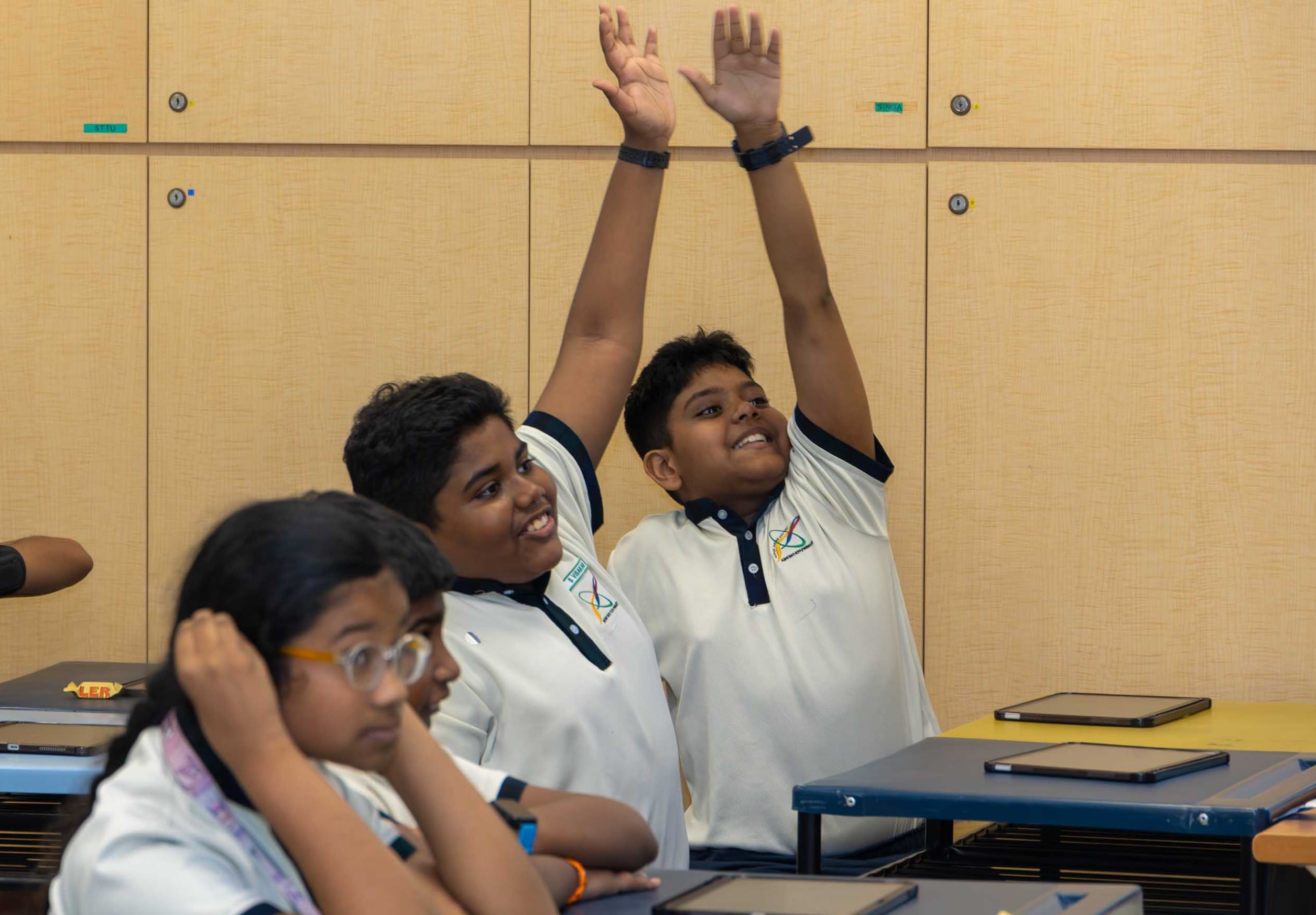 Two schoolboys in white uniforms raise both hands in class; other students and tablets on desks in front.