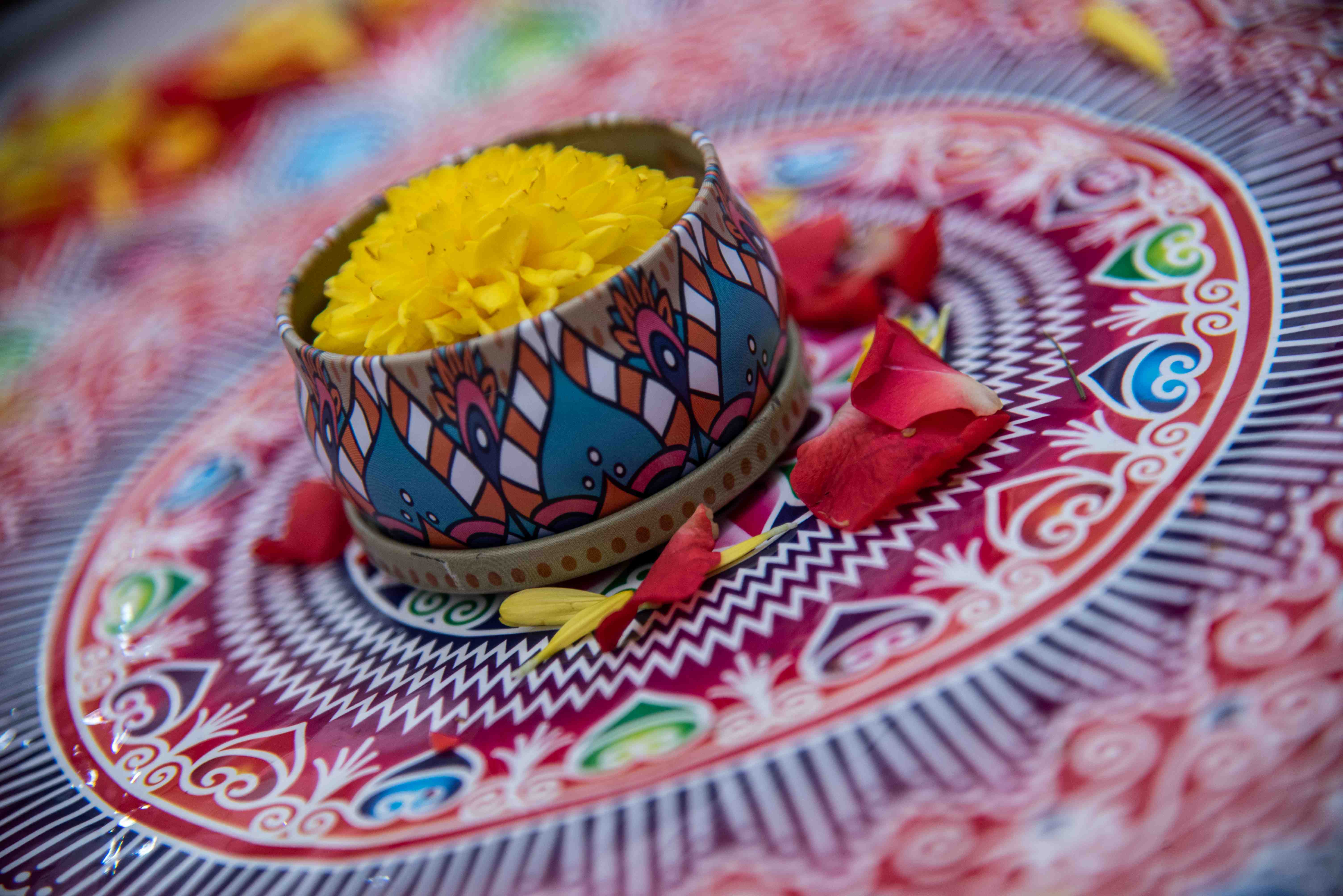 Patterned bowl filled with yellow flower petals on a colorful plate, with scattered red rose petals.
