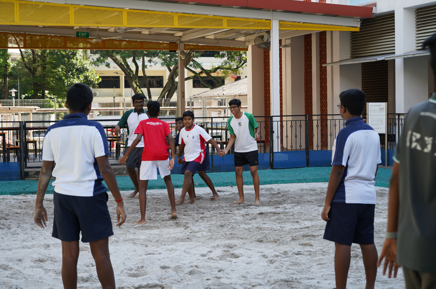 Students play a barefoot game on a sandy court under a roof, surrounded by school buildings.