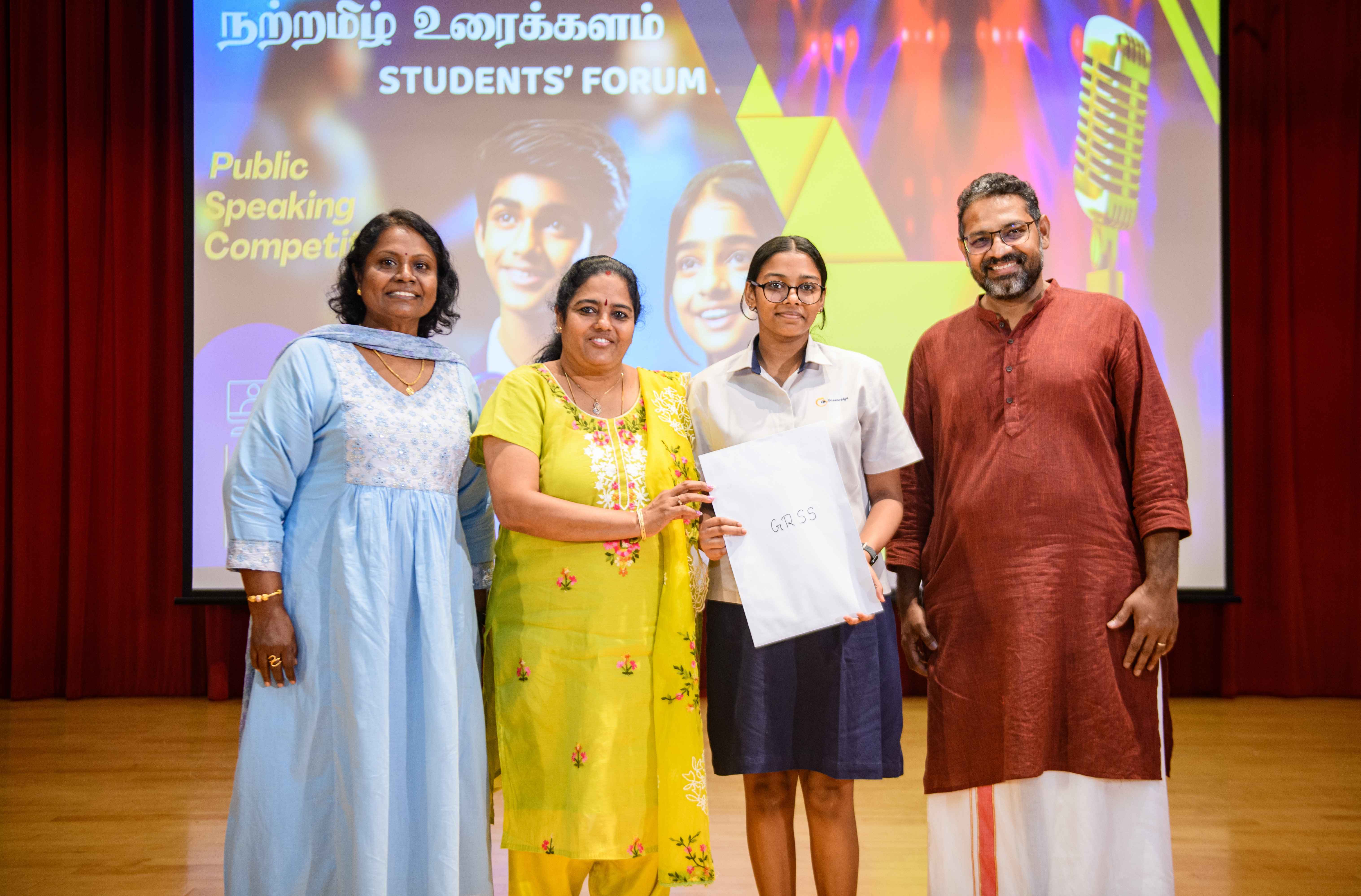 Four people pose on stage as a student holds a certificate; banner behind reads Students' Forum Public Speaking.