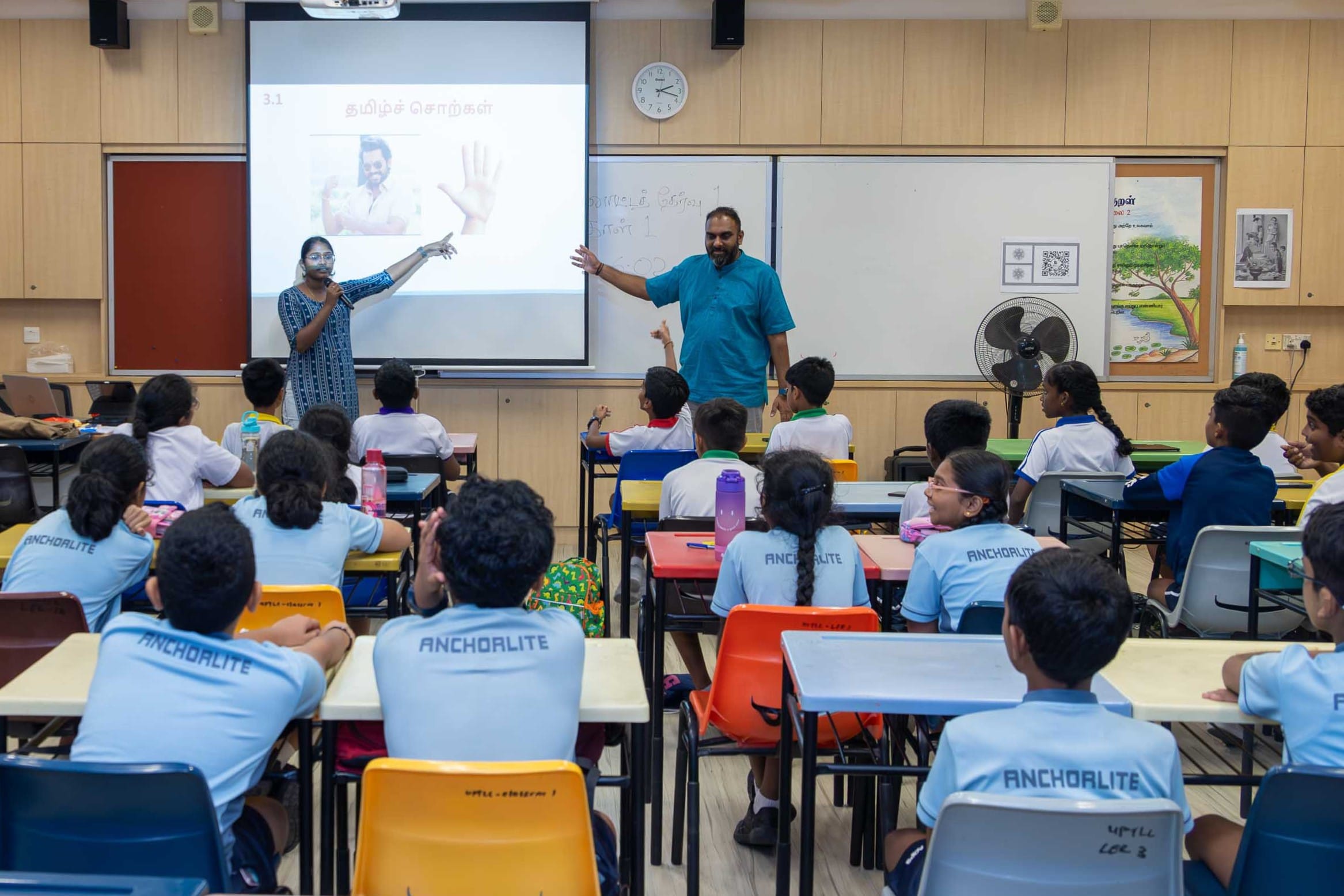Classroom with students seated as two teachers stand by a projector screen, pointing while students raise hands.