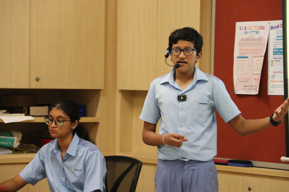 Student in blue uniform speaks with headset mic, gesturing, while another student sits at a desk nearby.
