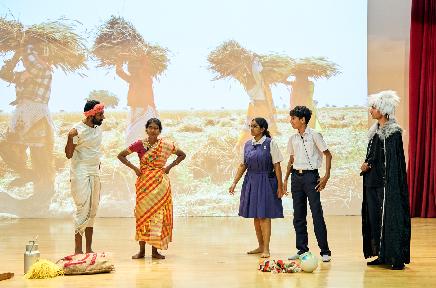 Five performers onstage before a projected farm scene; two in school uniforms, one in sari, one in cloak.