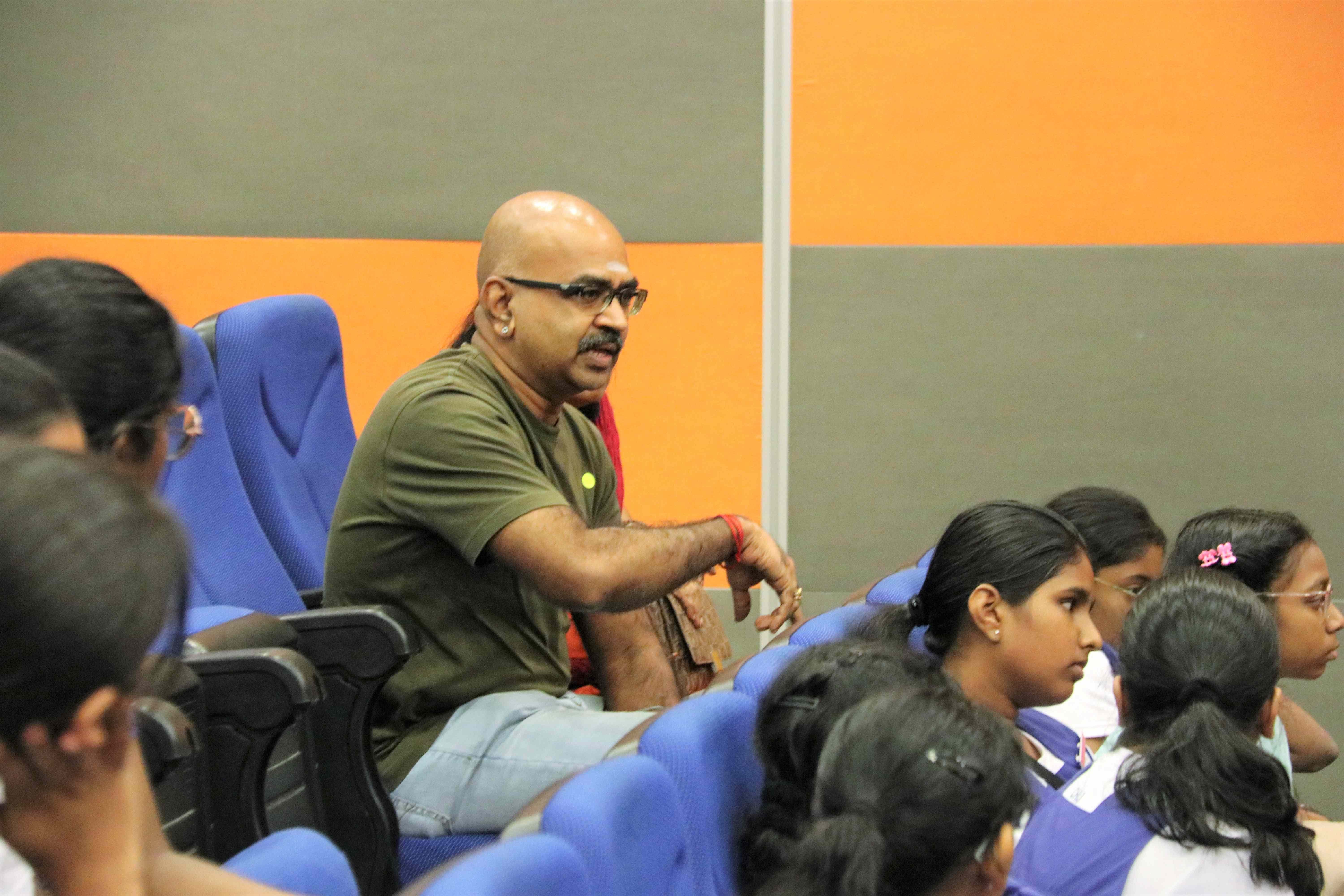 Man with glasses speaks while seated among students in blue auditorium chairs against orange and gray walls.