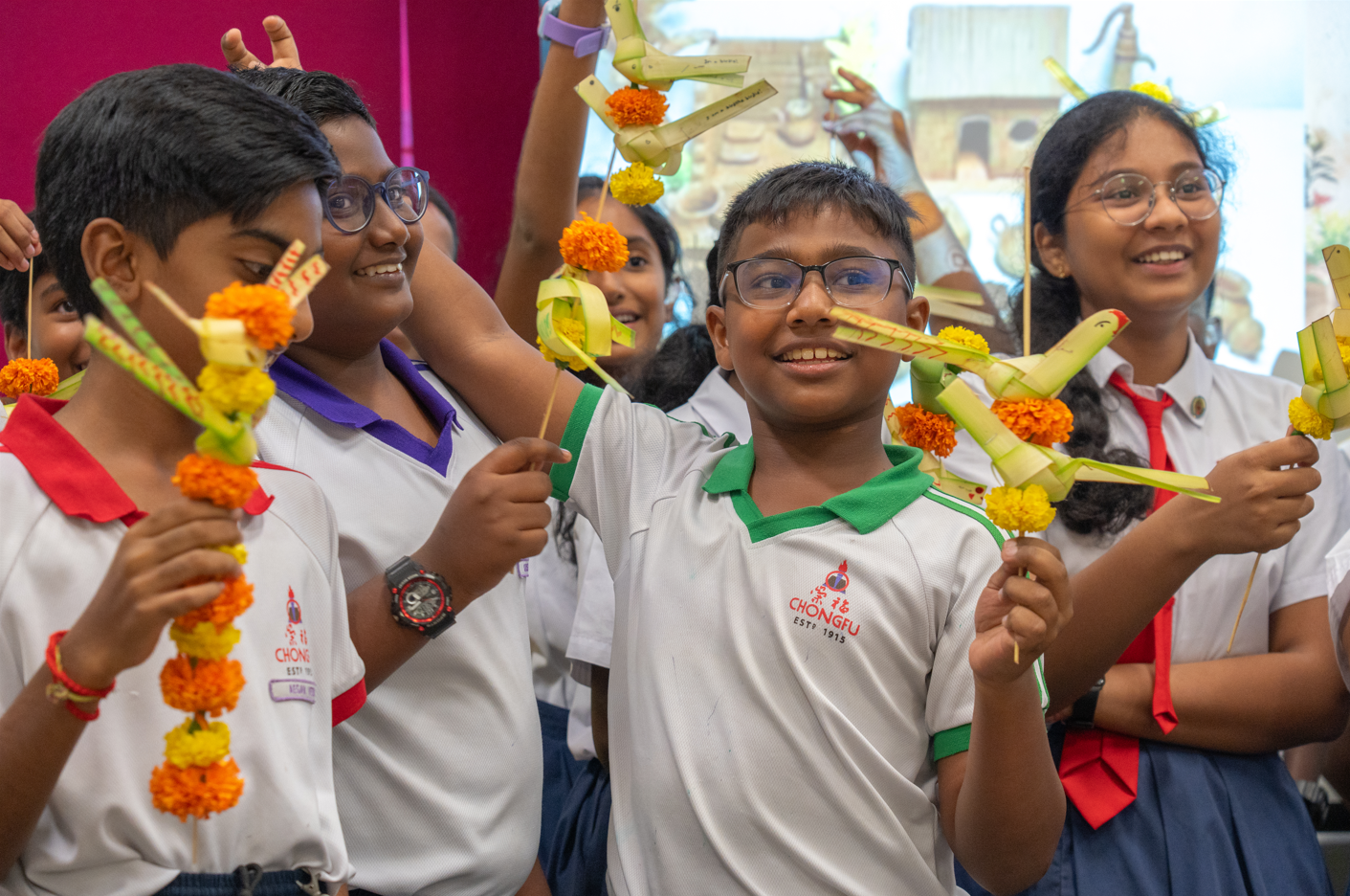 Smiling schoolchildren hold palm-leaf toys decorated with marigold garlands in a classroom.