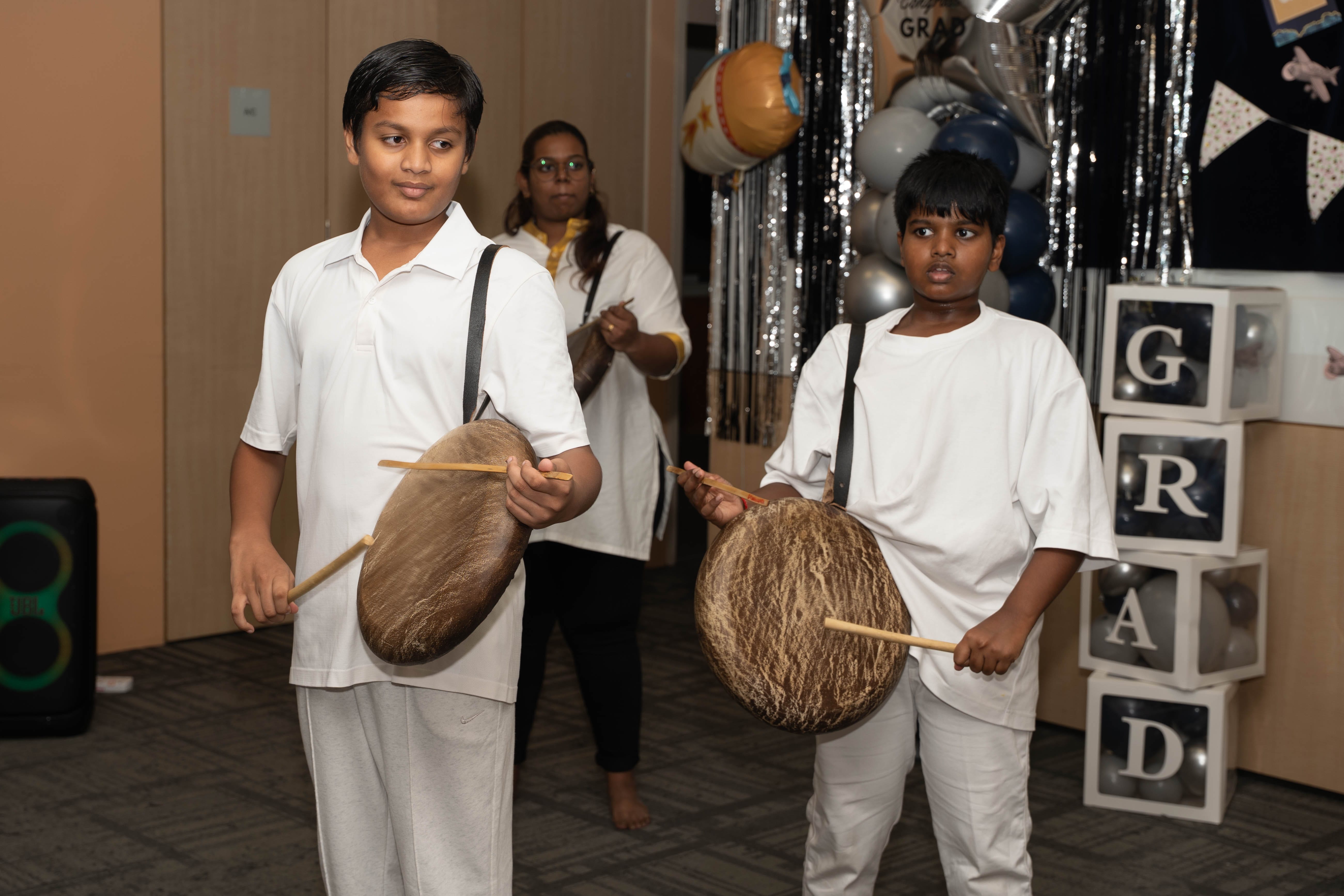 Two boys in white outfits hold drums and sticks at a party backdrop with balloons and letter blocks.