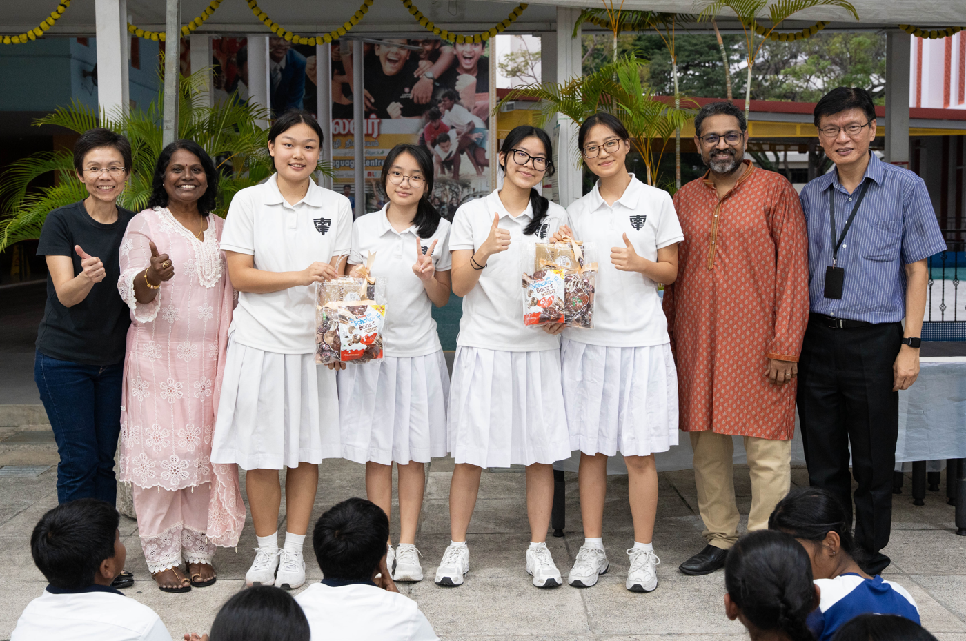 Six students in white uniforms pose with three adults, holding gift baskets and giving thumbs up.