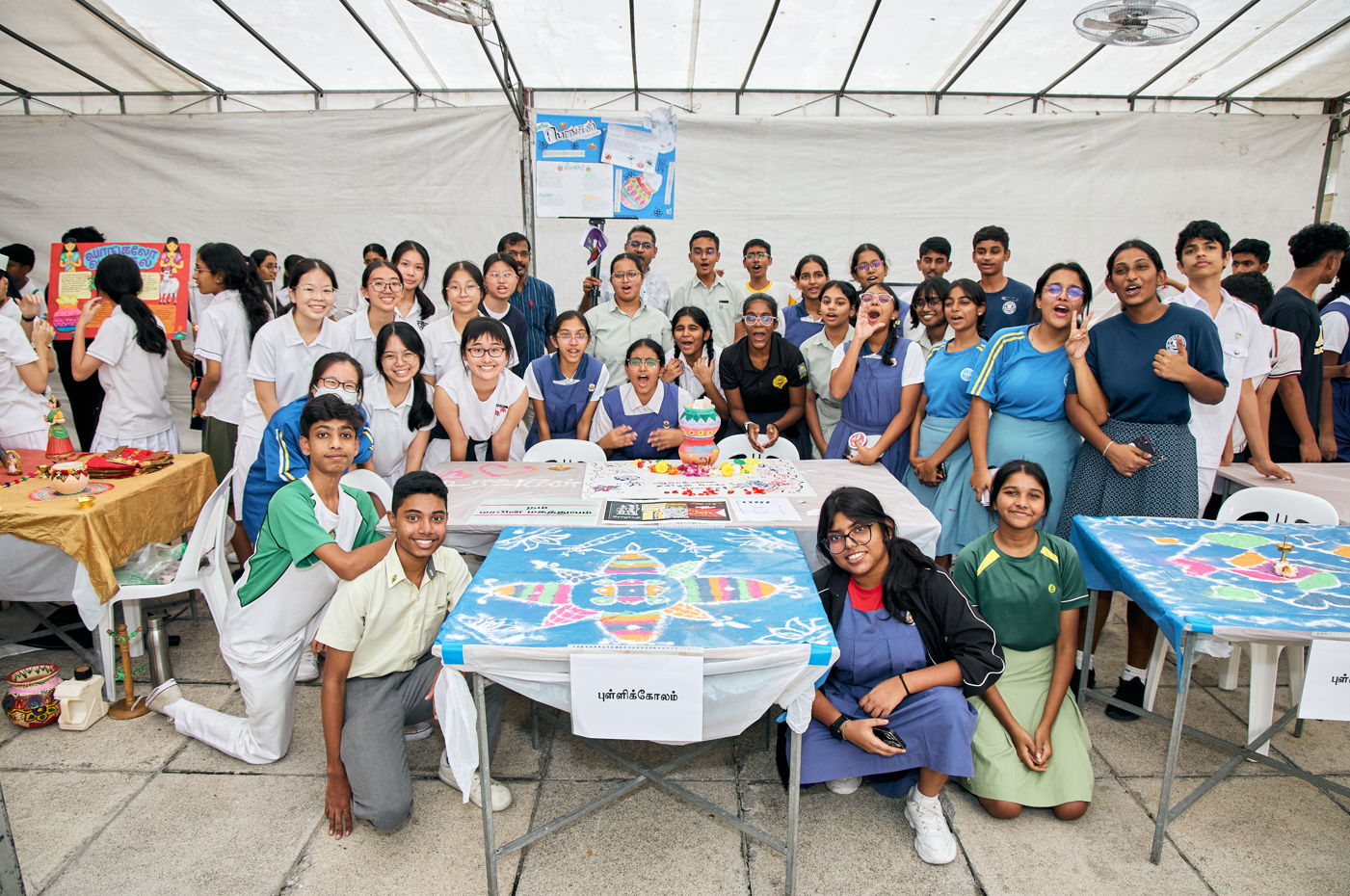 Group of students posing in a tent behind tables displaying colorful rangoli-style artworks and crafts.