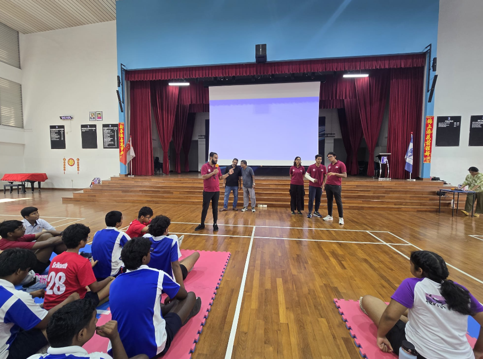 Students sit on mats in a gym as five presenters stand before a stage with red curtains and a screen.