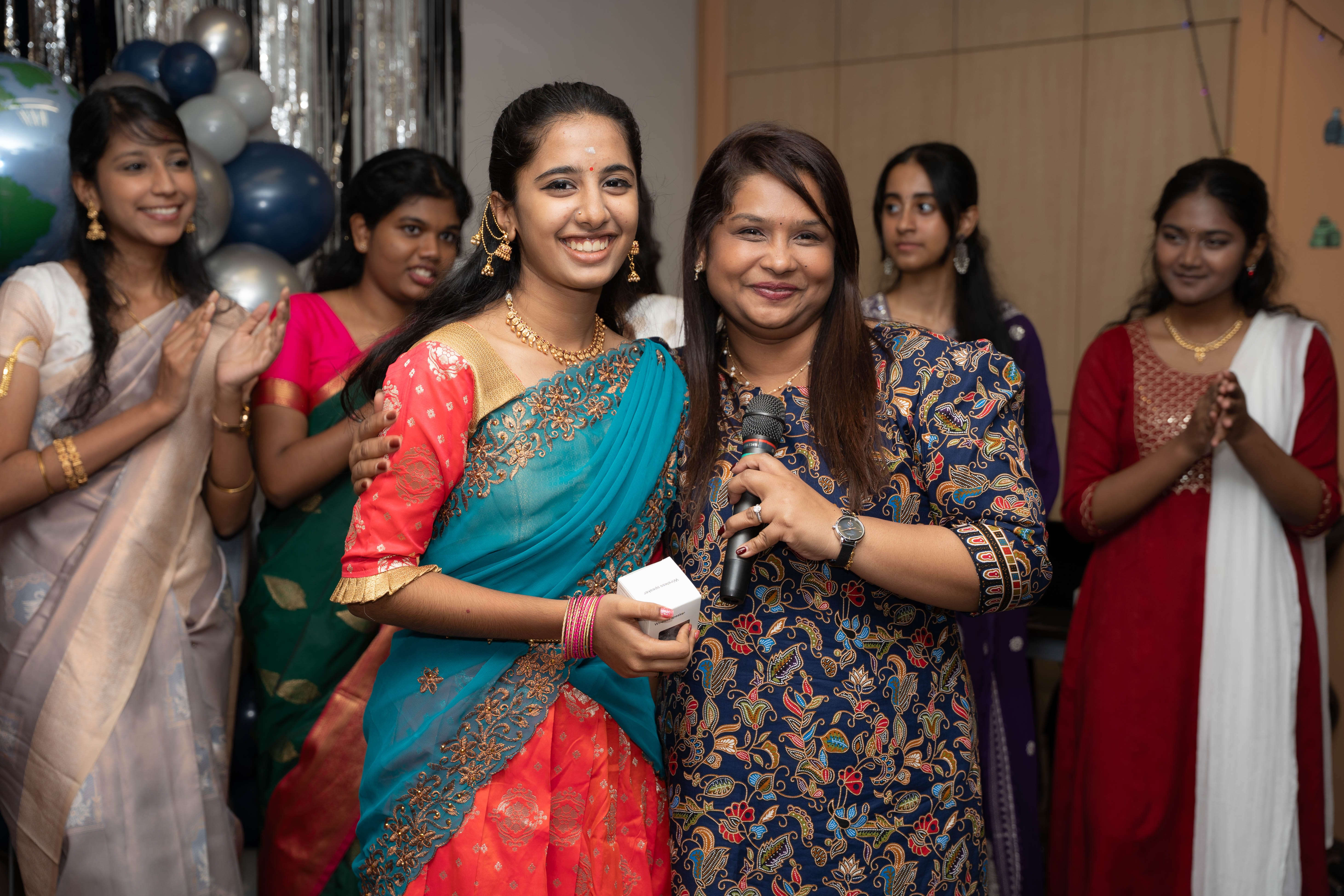 Two women smiling, one holding a microphone, while others in colorful dresses clap behind them.