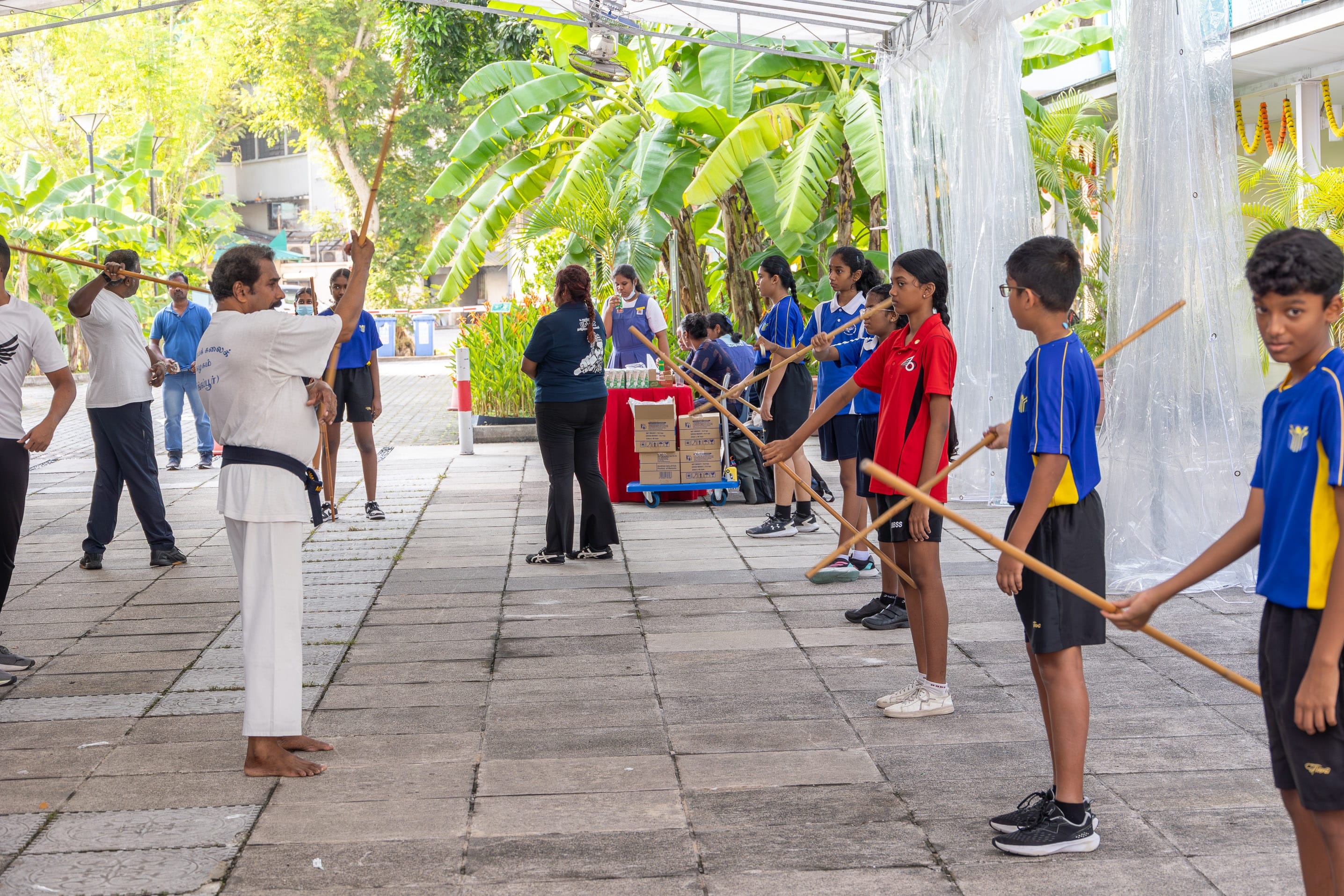 Instructor in white uniform leads kids holding wooden staffs in a line on a walkway with banana trees.