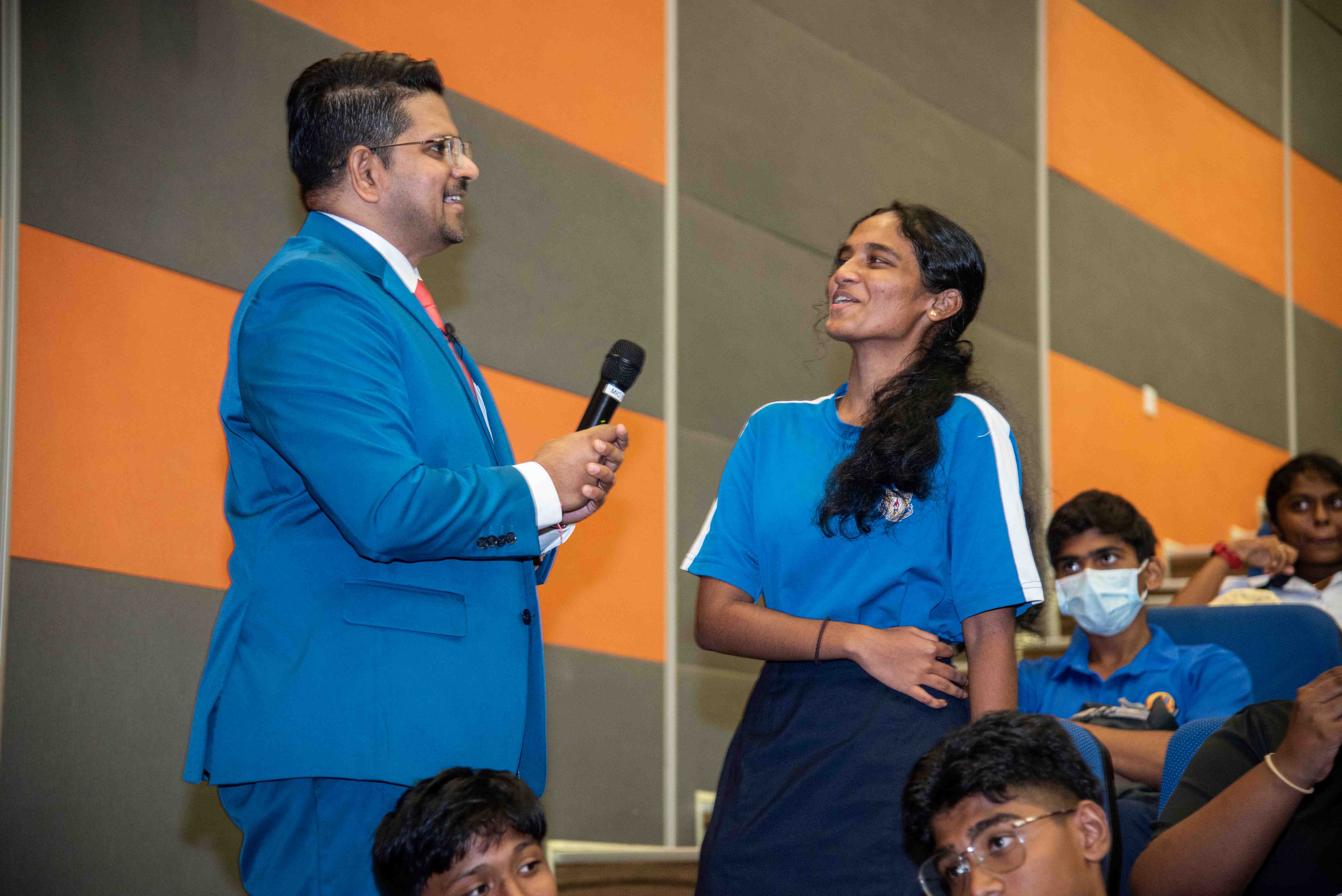 Man in blue suit holds a microphone toward a smiling woman in blue shirt, with seated audience behind.