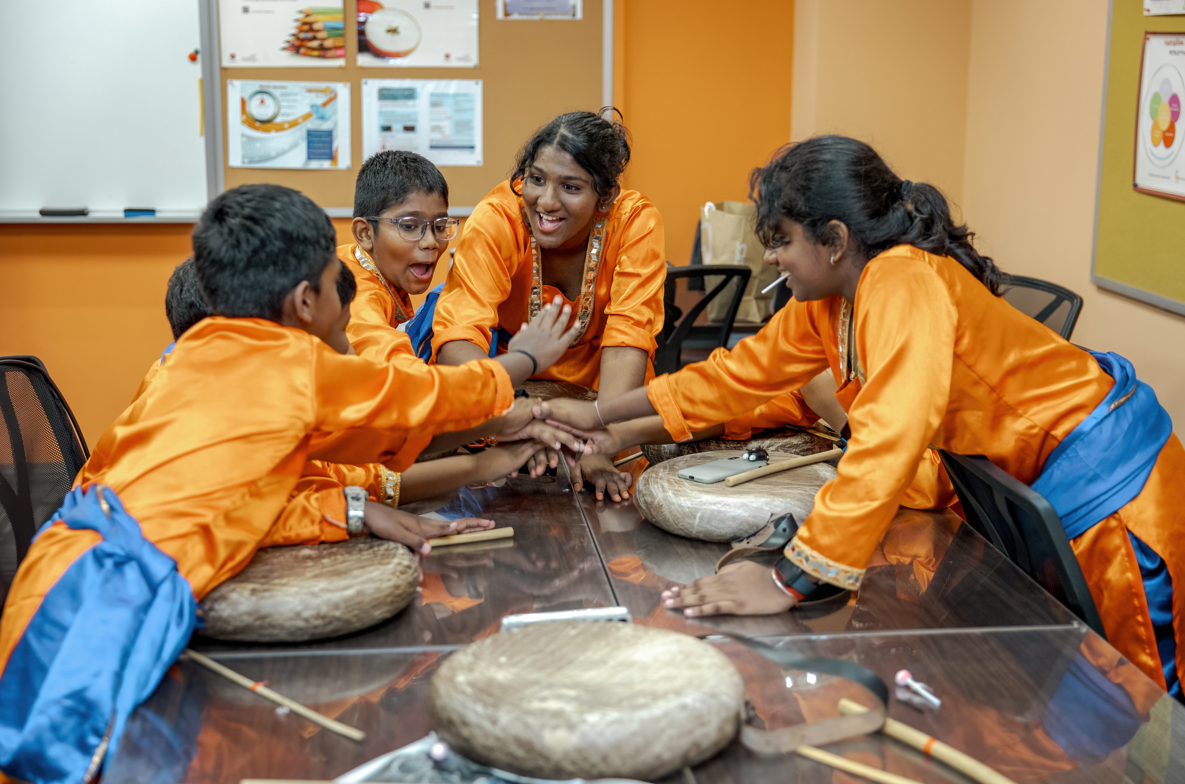 Five youths in orange outfits stack hands on a table with drums and sticks, smiling in a classroom.