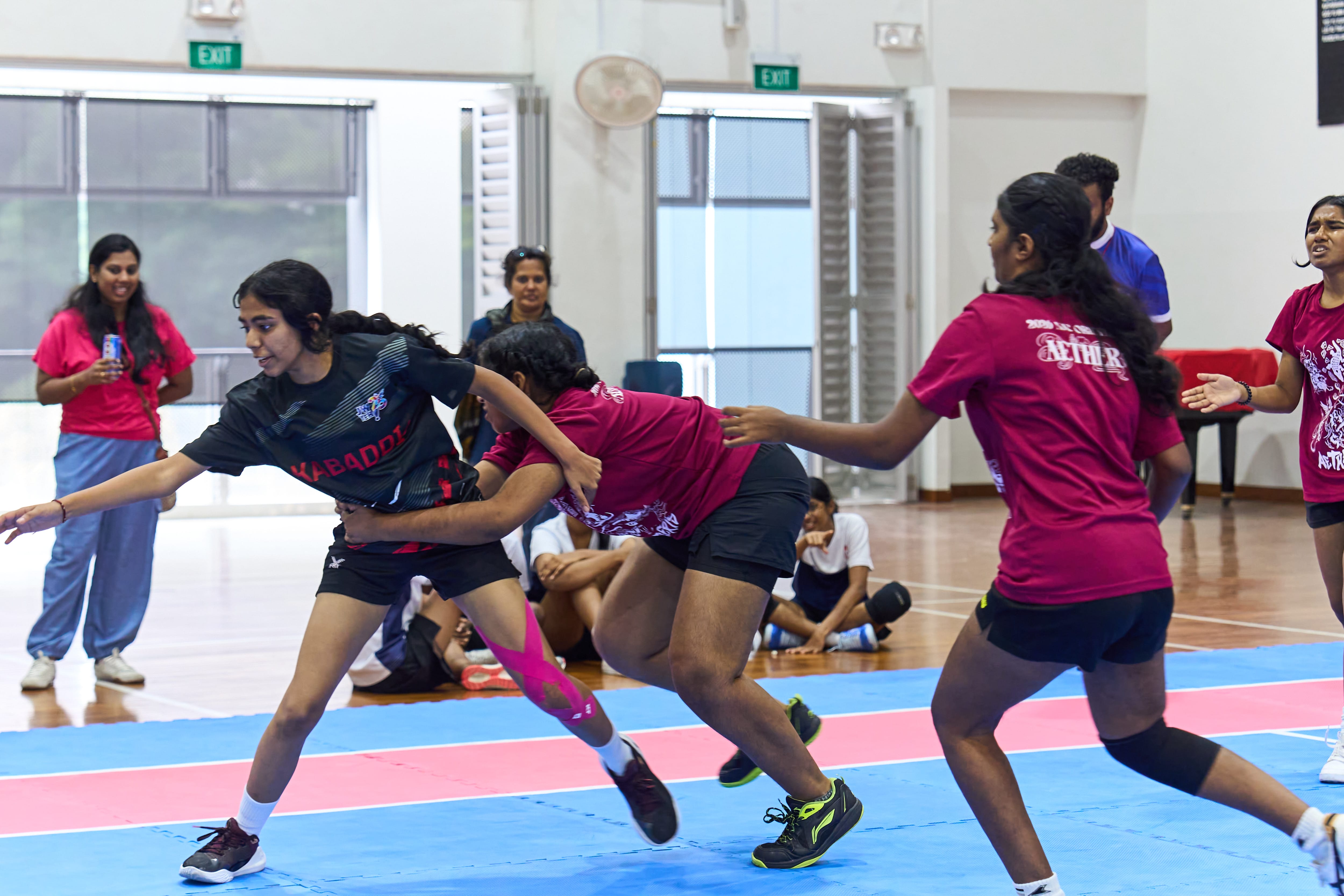 Indoor kabaddi match as a player in black is grabbed at the waist by a defender in maroon while reaching forward.