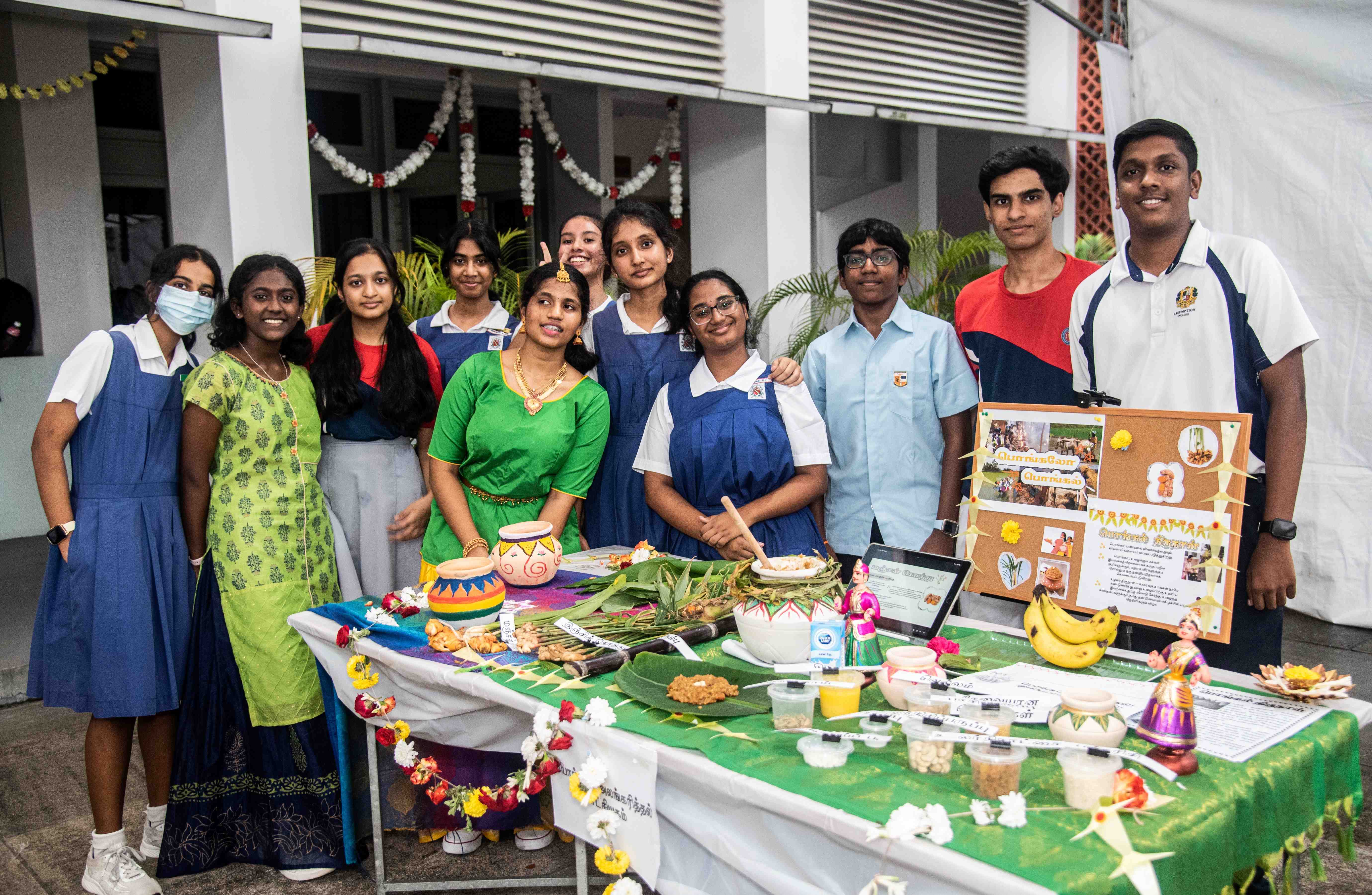 Group of students behind a decorated table with food items, pots, bananas, and a display board.