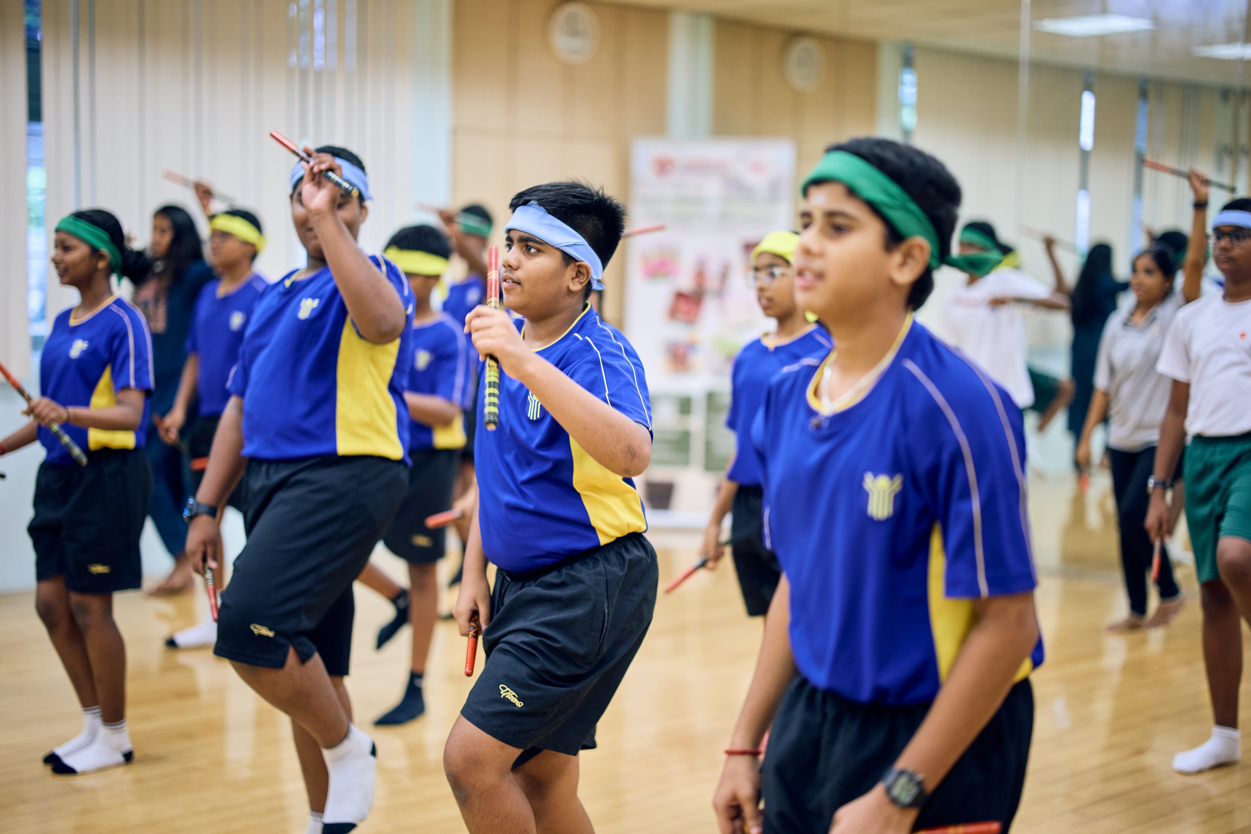 Students in blue jerseys and headbands practice a stick drill in a gym with mirrored walls.