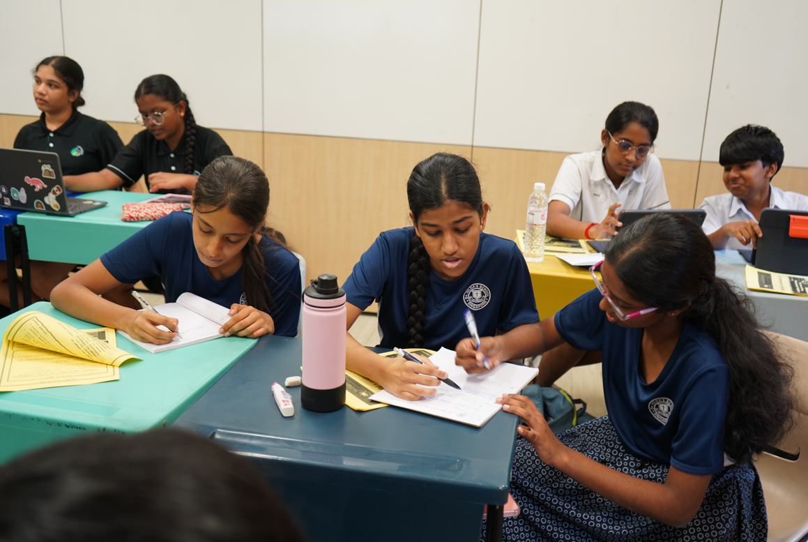 Students sit at classroom tables, writing in notebooks; one uses a laptop and others share notes.