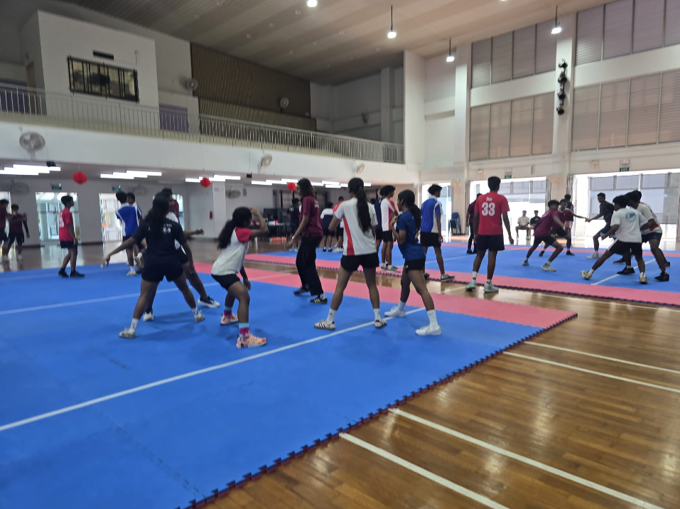 Students practicing kabaddi on blue mats in an indoor gym, with teams facing off and others watching.