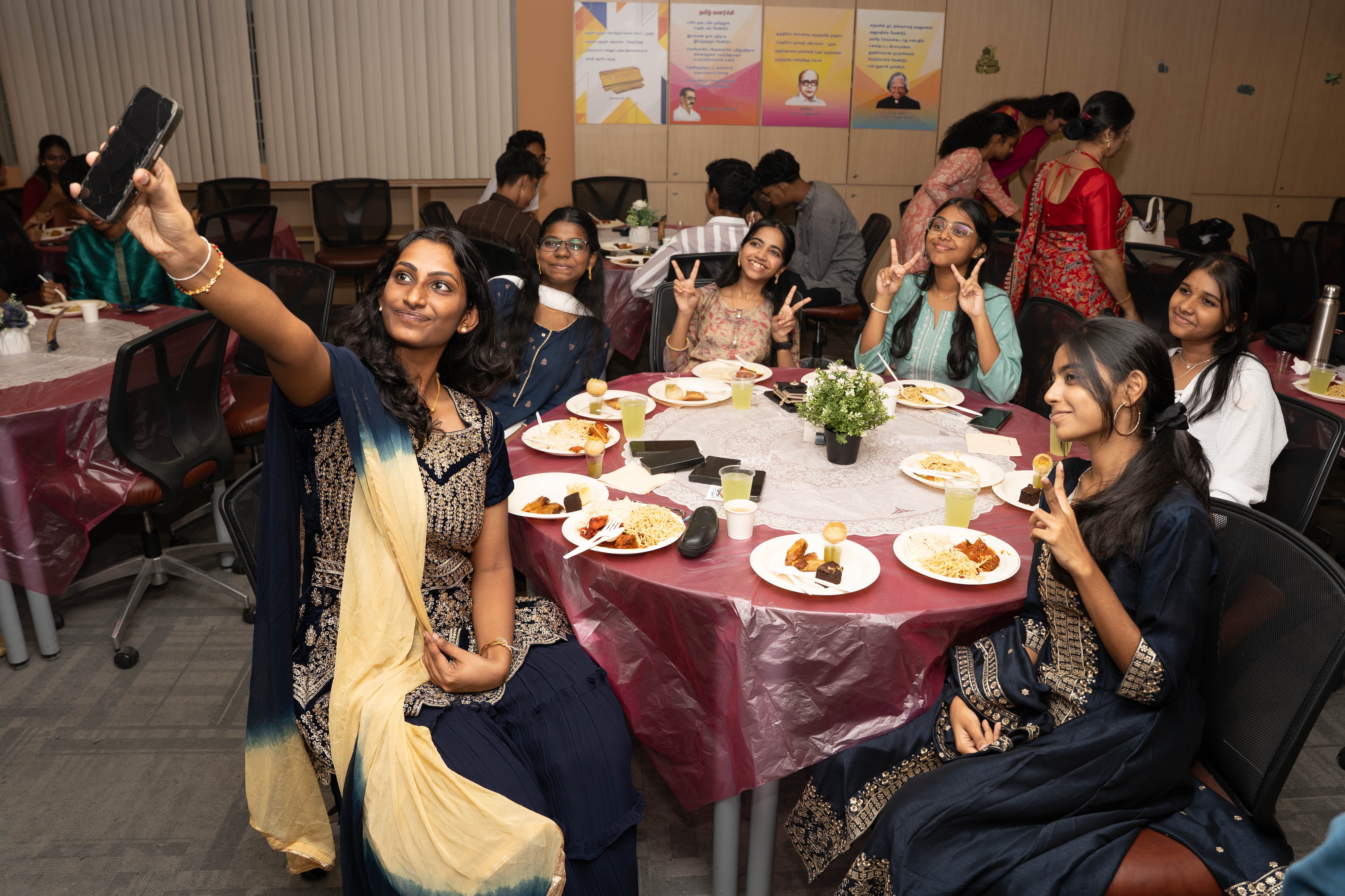 Woman takes a selfie as five friends pose with peace signs around a table with plates of food.