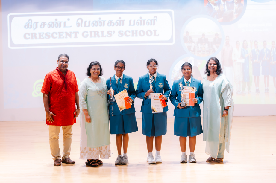Three schoolgirls in blue uniforms hold trophies and certificates, standing with three adults on stage.