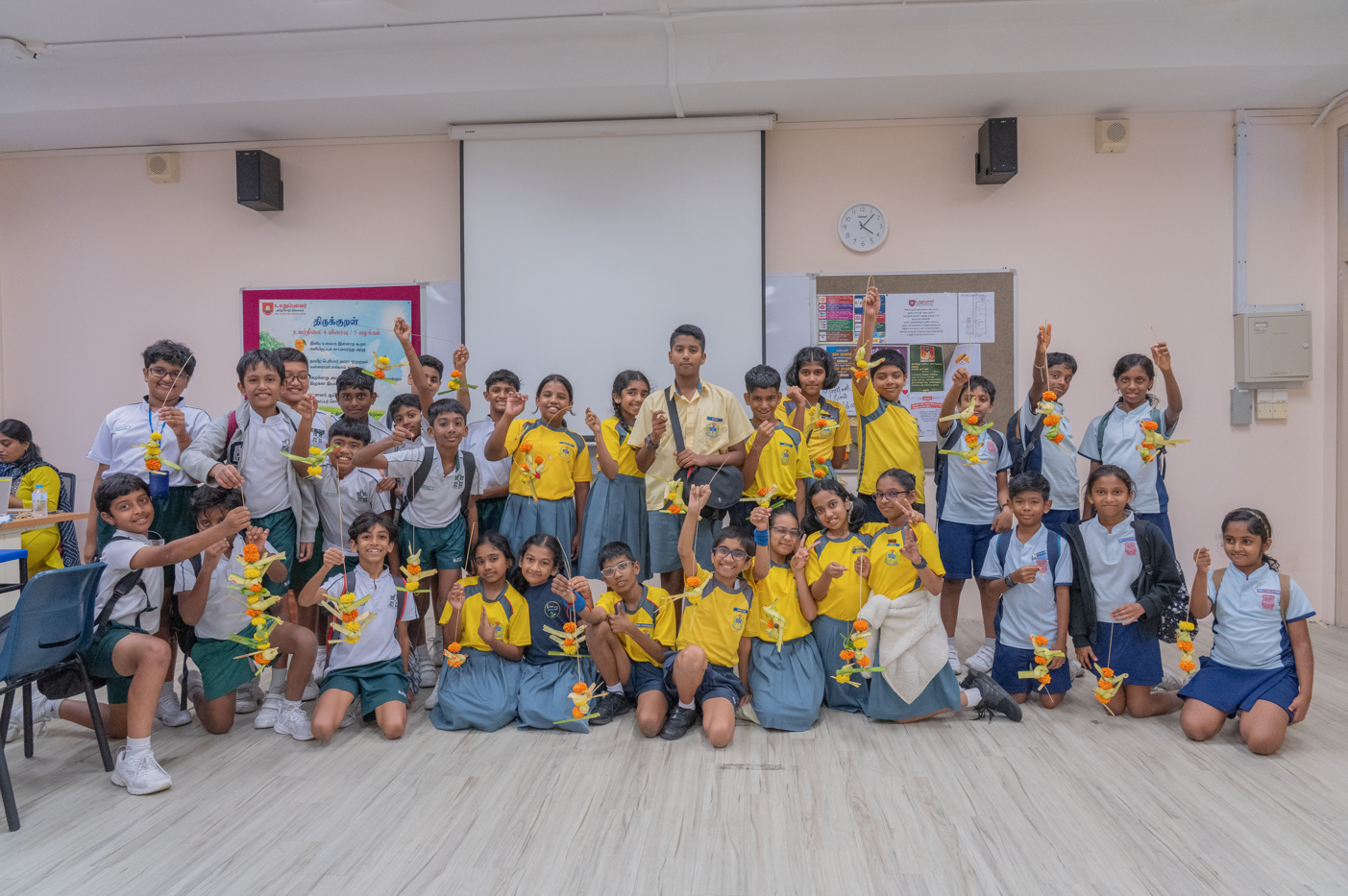 Group of schoolchildren in a classroom holding up flower garlands, some kneeling and some standing.