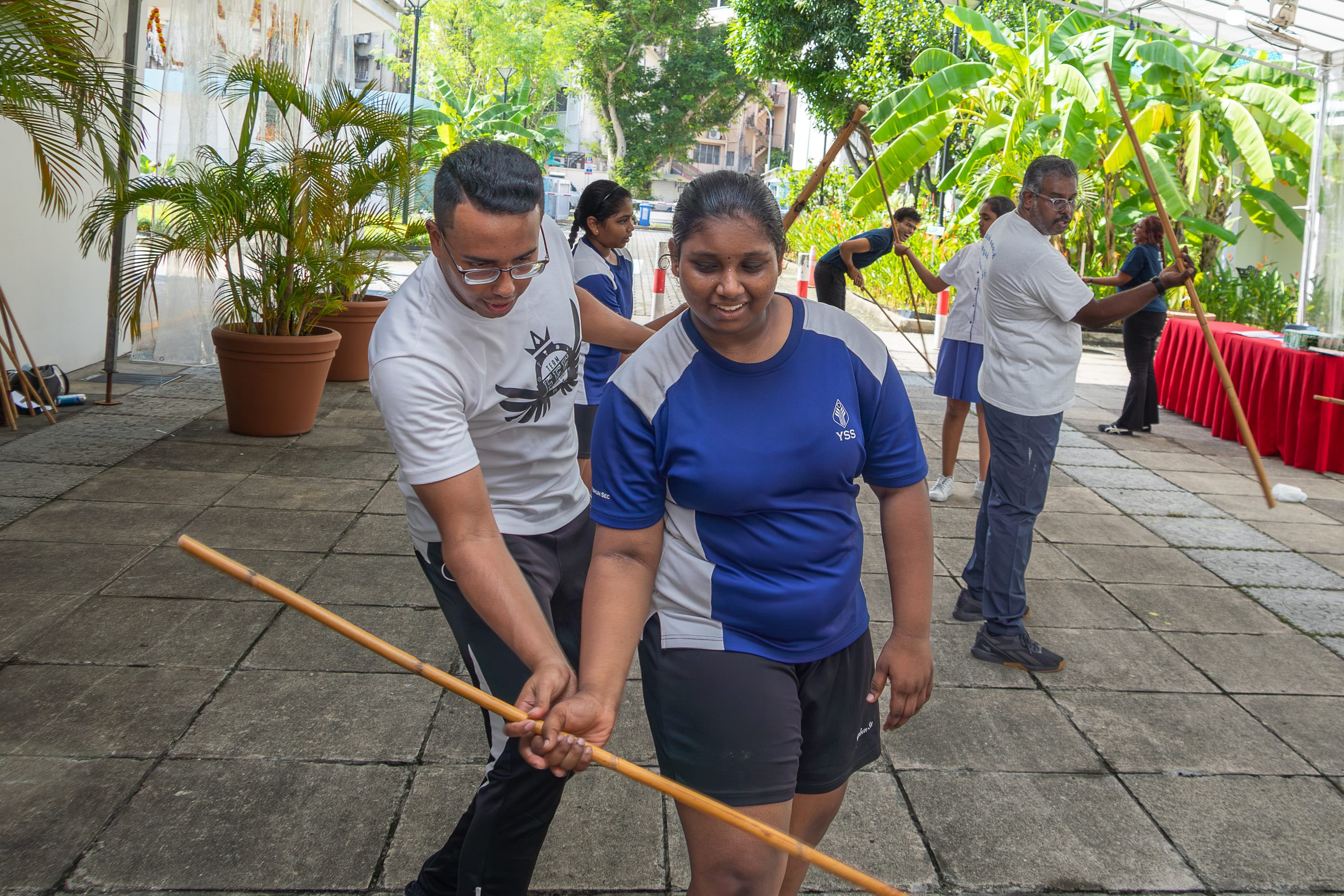 Man guides a girl holding a long wooden staff while others practice in an outdoor courtyard with plants.