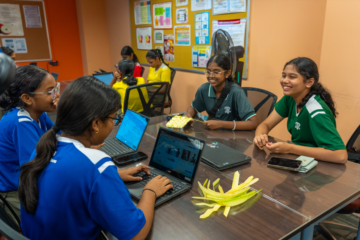Teens in school uniforms sit around a table with laptops and phones, chatting in a classroom.