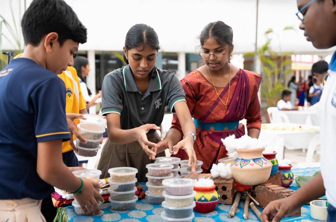 Students gather at a table, arranging stacked tubs of grains beside decorated pots and bricks.