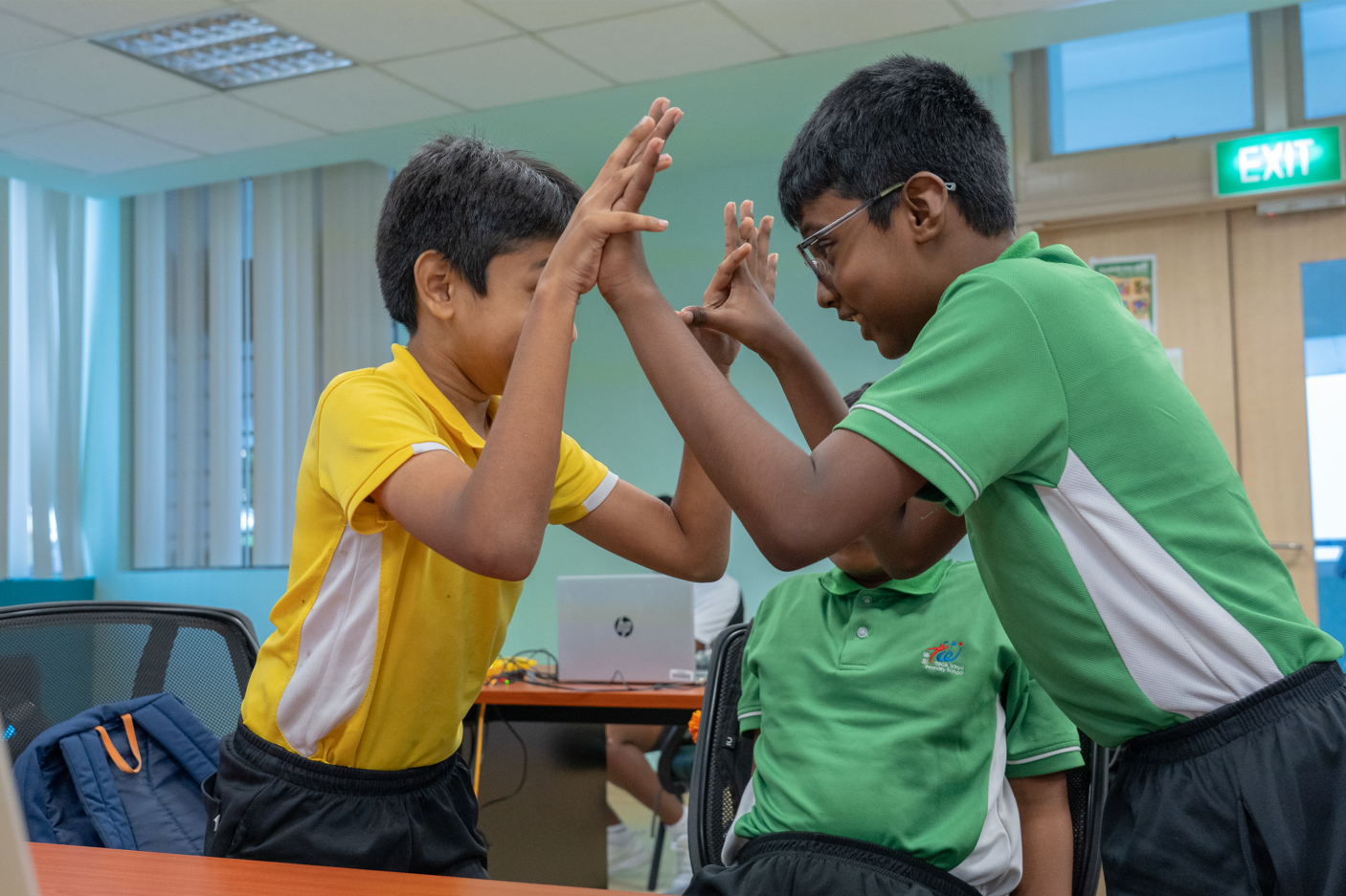 Two boys in yellow and green shirts face each other, hands raised as if playing patty-cake in a classroom.