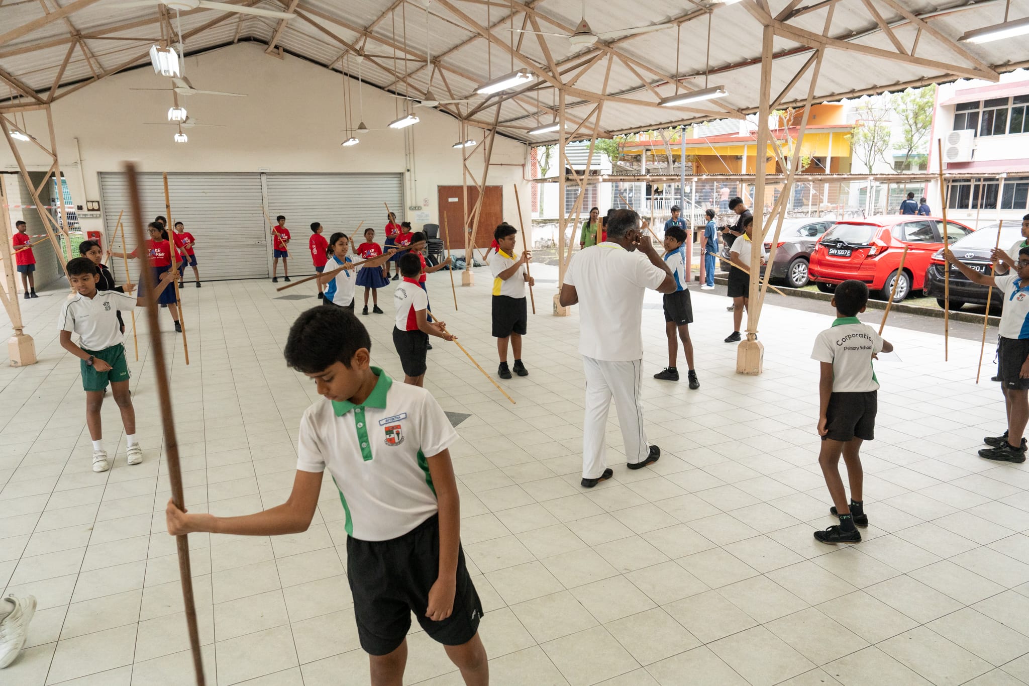 Schoolchildren practice with long wooden staffs in an open hall while an instructor in white watches.