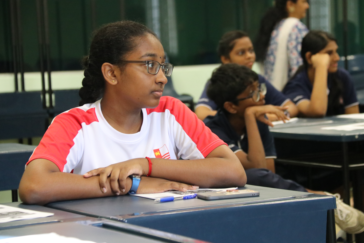Student with glasses in red-and-white shirt sits at desk, others blurred in classroom behind.