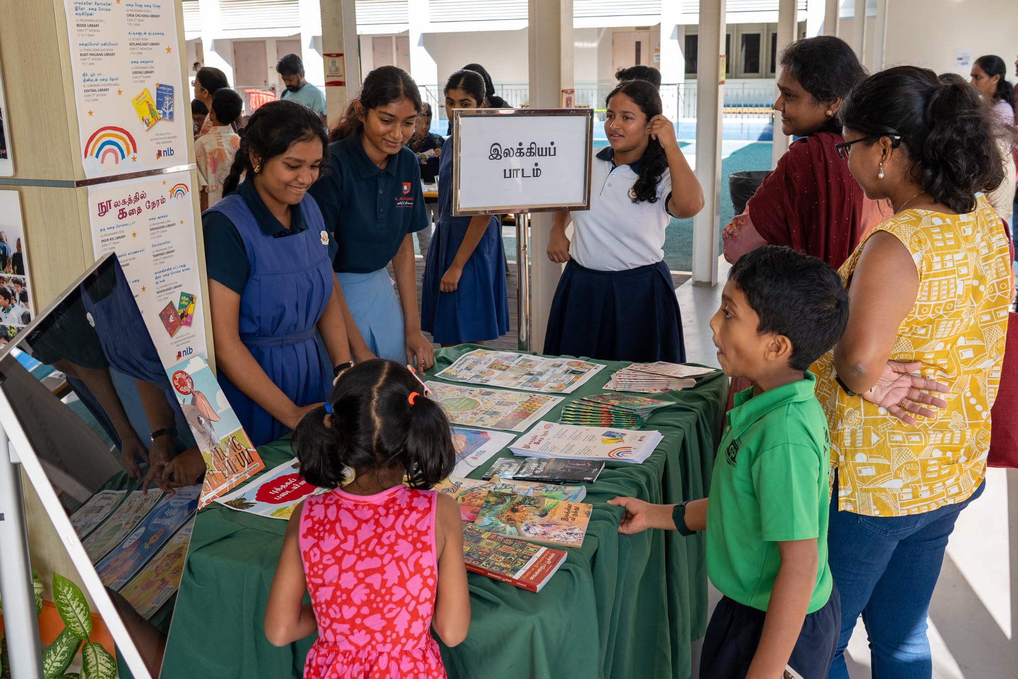 Students and adults browse books and posters at a table; Tamil sign on stand in a school corridor.