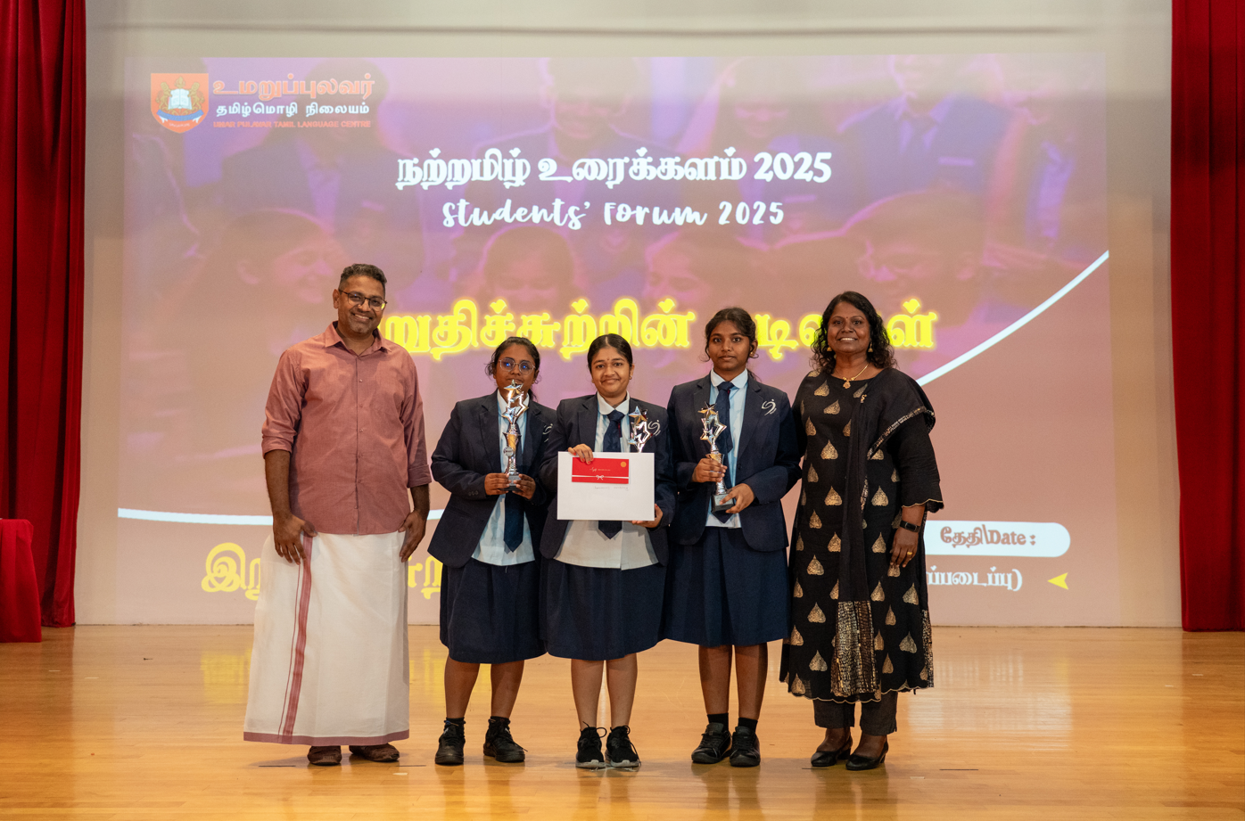 Five people on stage; three students in uniforms hold trophies and a certificate, flanked by two adults.