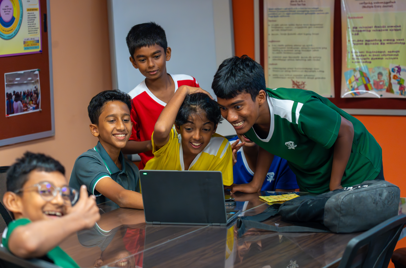 Five boys gather around a laptop at a table, smiling and leaning in, with posters on the wall.