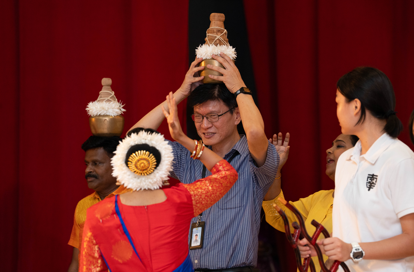 Man holds decorated pot on his head as a woman in red and blue dress raises her hands in front of him.