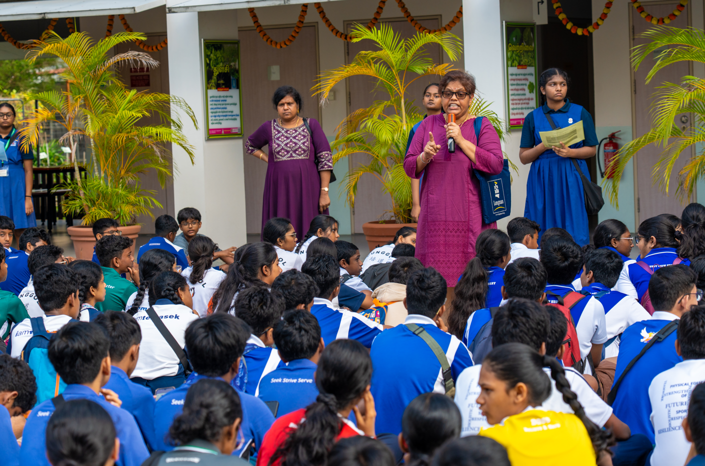 Woman in magenta speaks into a microphone to seated students in uniforms in a courtyard with potted palms.