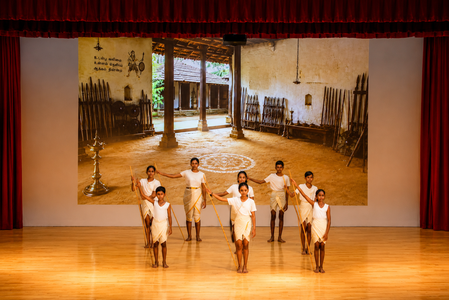 Eight performers in white hold long staffs on a stage, before a large courtyard scene backdrop framed by red curtains